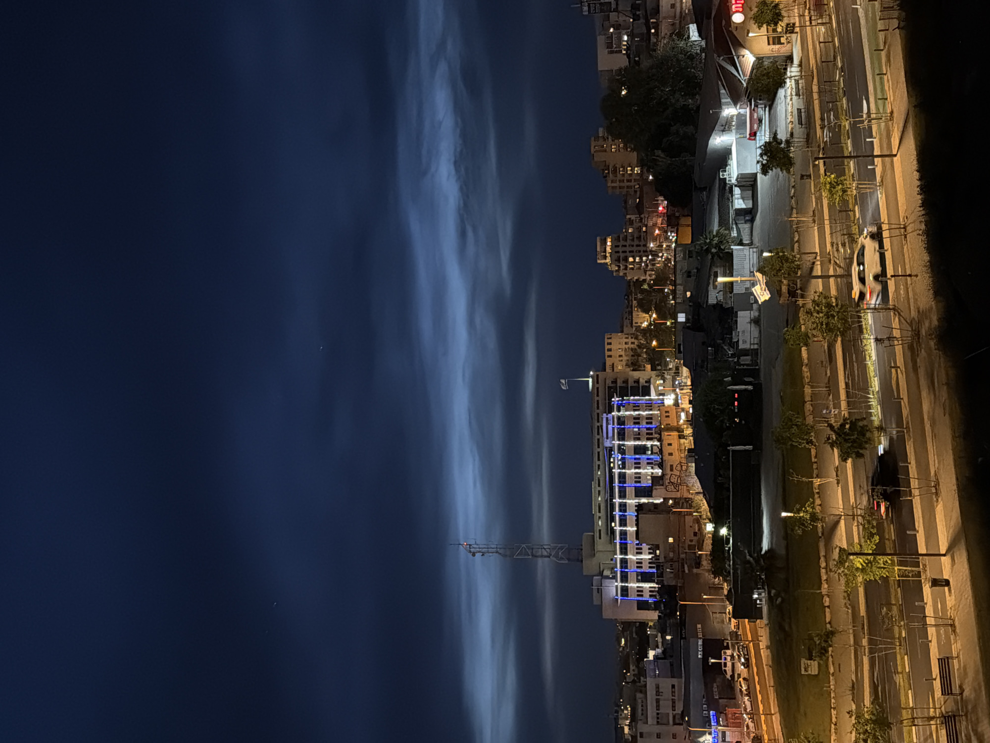 A panoramic nighttime photograph captures the vibrant cityscape of Tel Aviv, Israel. The dark blue sky occupies the upper portion of the frame, featuring long, streaky, light-colored clouds that span horizontally across the expanse, suggesting a long exposure technique or unique illumination.
The city sprawls across the lower half of the image, showcasing numerous buildings aglow with artificial lights. A prominent building stands out with distinct blue and white decorative lights illuminating its facade. A tall, slender communication tower, tipped with a small red light, rises from the heart of the urban area.
In the foreground, a multi-lane road runs parallel to the buildings, brightly lit by warm yellow streetlights. Trees line the pavement, interspersed with various street furniture including benches and signs, some with indistinct text. A car, its headlights or taillights appearing blurred due to motion, is visible on the road. The scene depicts typical evening activity in a bustling urban environment under a dramatic sky. No people are clearly visible.