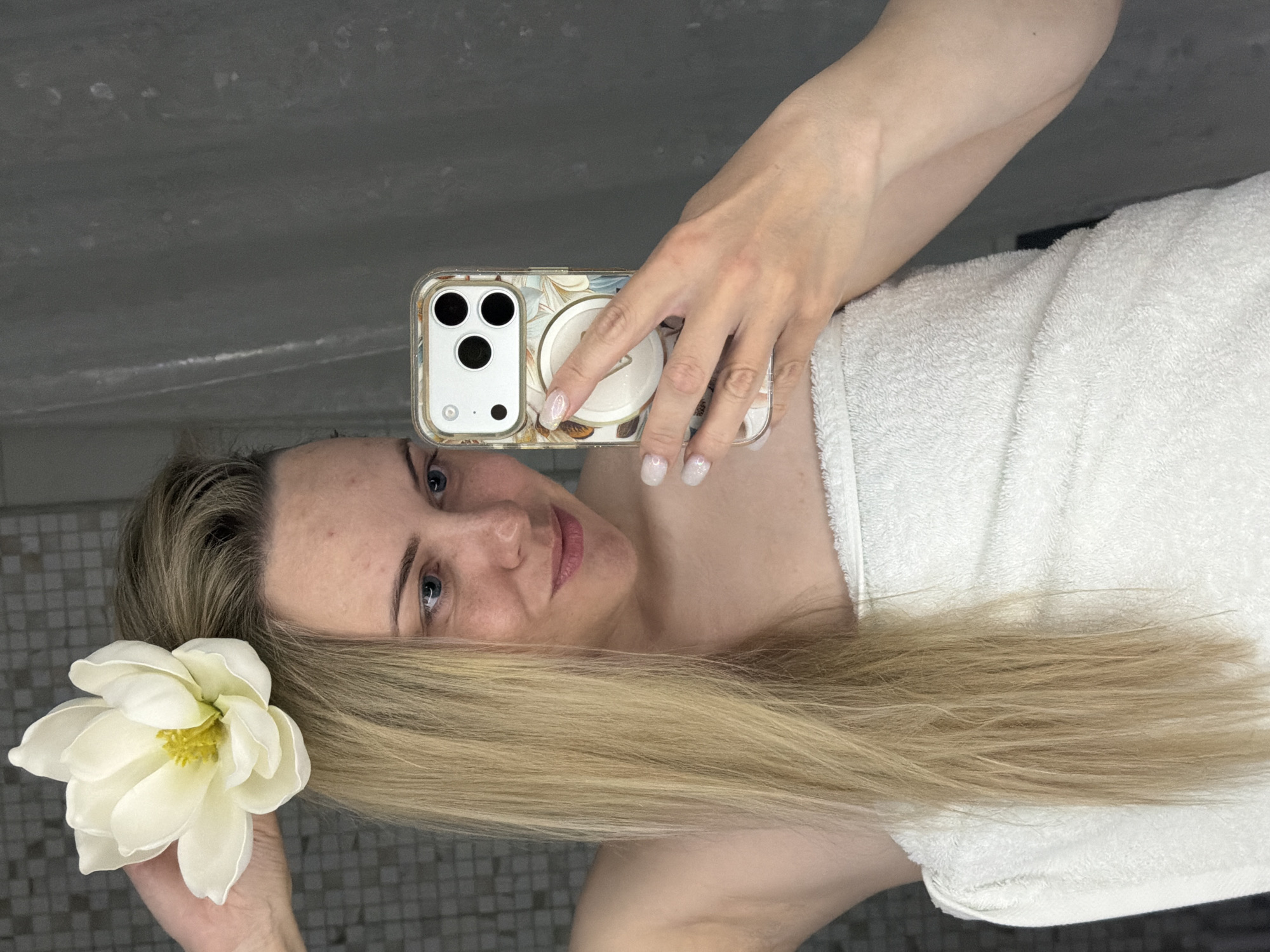 The image, rotated 90 degrees counter-clockwise, displays a woman taking a mirror selfie in an indoor setting, likely a bathroom or spa. The woman, with long, straight blonde hair and light blue eyes, is shown from the upper chest upwards, with her head oriented towards the left side of the frame in the current rotation. She has a gentle, subtle smile and looks directly into the camera.
A large, white artificial flower with a yellow center is tucked into her hair near her left ear. She is wrapped in a white terry cloth towel. Her right arm is raised, holding a white smartphone, which appears to be an iPhone with three camera lenses, encased in a clear cover featuring a delicate floral or abstract design. A white circular grip (pop socket) is attached to the back of the phone. Her fingernails are manicured with a light, shimmering polish, and a ring is visible on her ring finger.
The background reflected in the mirror shows a grey tiled wall behind her head and a plain, textured grey surface above it, consistent with a bathroom or similar area. The lighting is soft and even, characteristic of an indoor environment. The woman appears relaxed and content. No visible text is present in the image. The location context provided is Votkinsk, Russia.