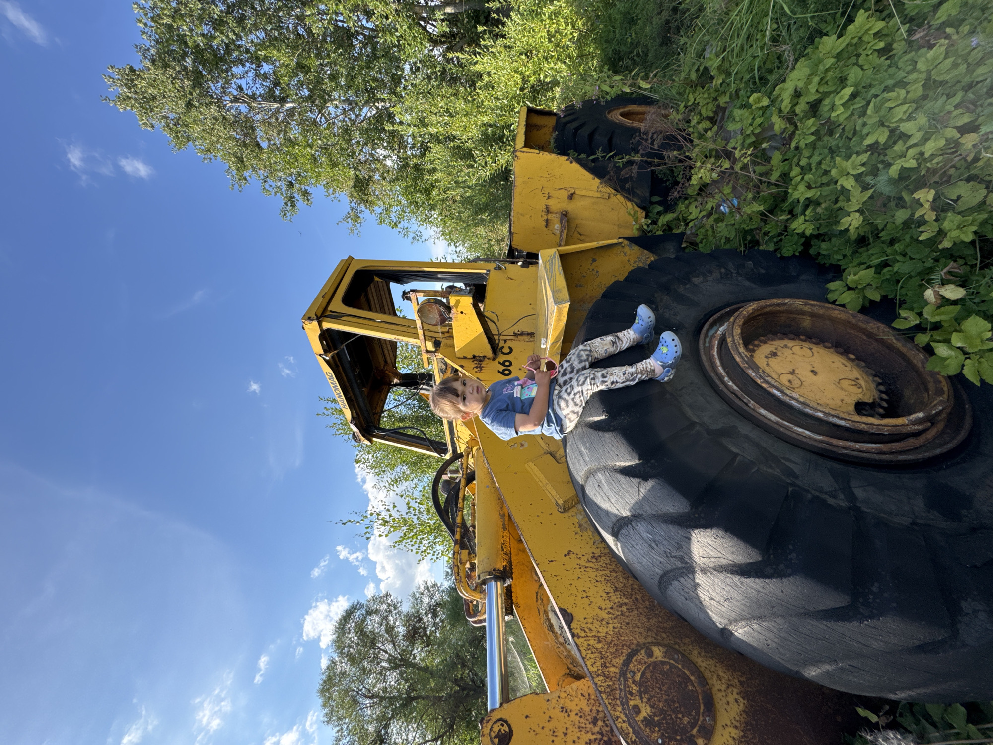 Innolovo, Russia. A young child, dressed in a blue t-shirt and patterned leggings, sits on the massive tire of a yellow, weathered construction vehicle, possibly a loader. The vehicle appears to be stationary and partially overgrown with greenery. The child is holding a pink object. The background consists of trees and a bright blue sky with scattered clouds. The scene suggests an outdoor, possibly rural or abandoned, setting.