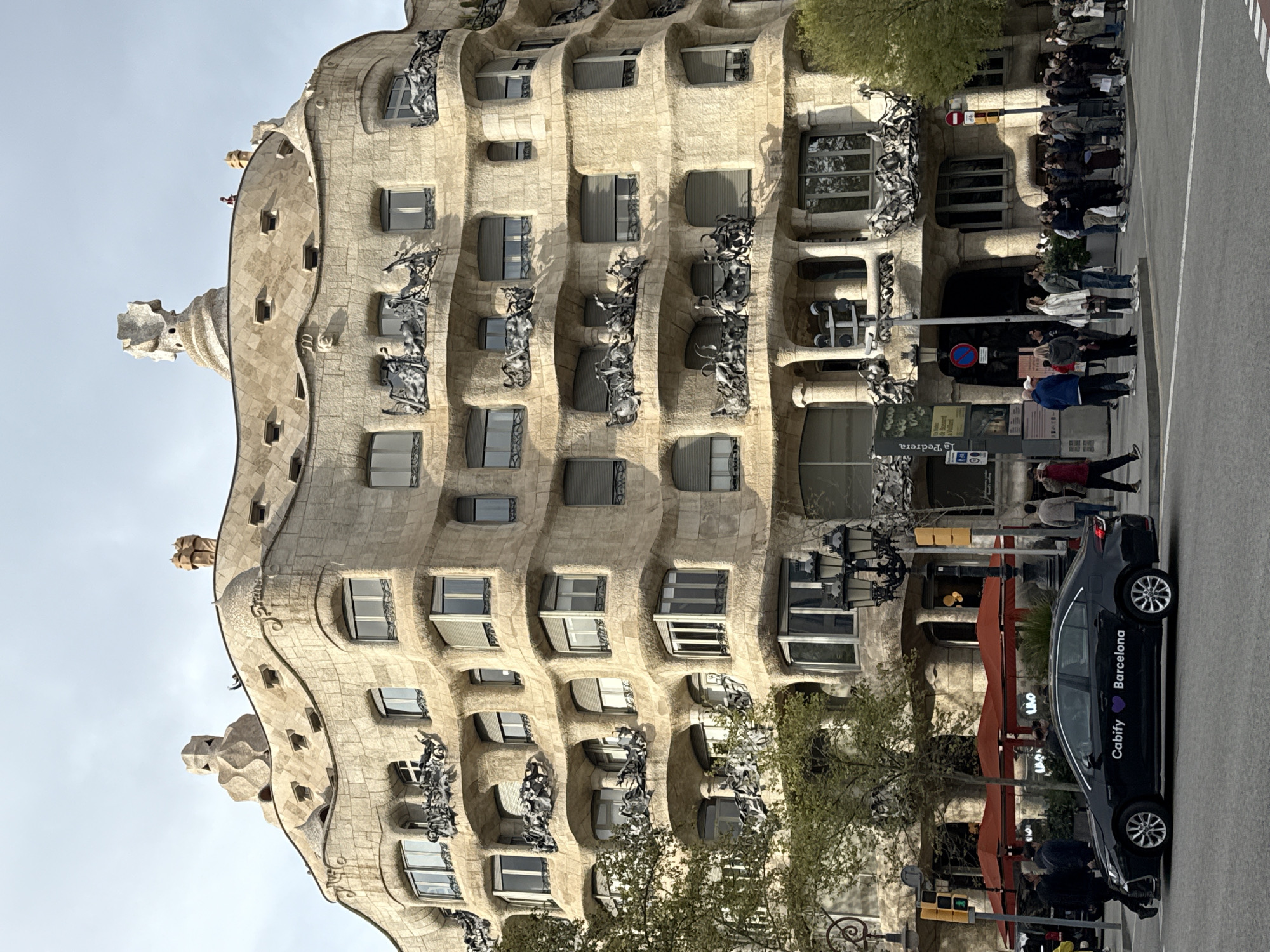 The image depicts Casa Milà, also known as La Pedrera, a modernist building in Barcelona, Spain, designed by Antoni Gaudí. The structure, noted for its undulating stone facade and sculptural iron balconies, is a prominent architectural landmark. A crowd of people stands along the sidewalk in front of the building, seemingly admiring or preparing to enter. A dark-colored car with the words "Cabify Barcelona" written on the side is parked along the street adjacent to the building, suggesting it may be a taxi or ride-sharing vehicle. Traffic lights and pedestrian signs are visible, adding to the urban context. The sky is lightly clouded, indicating daytime.