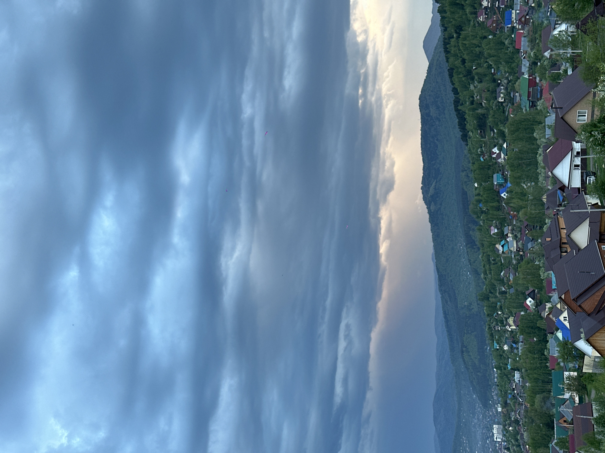 The image shows a panorama in Gorno-Altaysk, Russia, with a cloudy sky dominating the left half. The right half features a mountain covered in green forest. In the foreground, a residential area with closely packed houses is visible; the roofs of the houses display a variety of colors and materials. The image appears to be taken from an elevated position, overlooking the landscape.