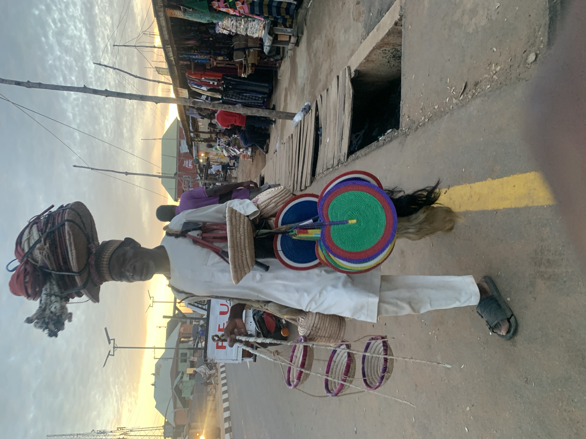 The media file shows a street vendor in Akwanga, Nigeria, at what appears to be sunset. The sky is filled with clouds and the golden light suggests the sun is low on the horizon. The vendor, a person of color wearing a white outfit and a distinctive hat with woven decorations and bundles on top, is the central subject. They are holding a string of woven baskets. Several colorful, circular fans, likely part of their merchandise, are also visible.
The setting is a bustling market scene, with stalls of various goods, including textiles, visible in the background. The ground is dusty and uneven, with a visible yellow line painted on it. There is a drainage ditch with wooden planks beside the vendor. Other people are present in the background, some browsing stalls and others walking. The overall mood is active, with the setting sun casting a warm glow over the scene. One of the items carried by the vendor has "REU" written on it, possibly part of a larger word or logo. The vendor's facial expression is neutral, but the overall scene suggests a typical day in a Nigerian market.