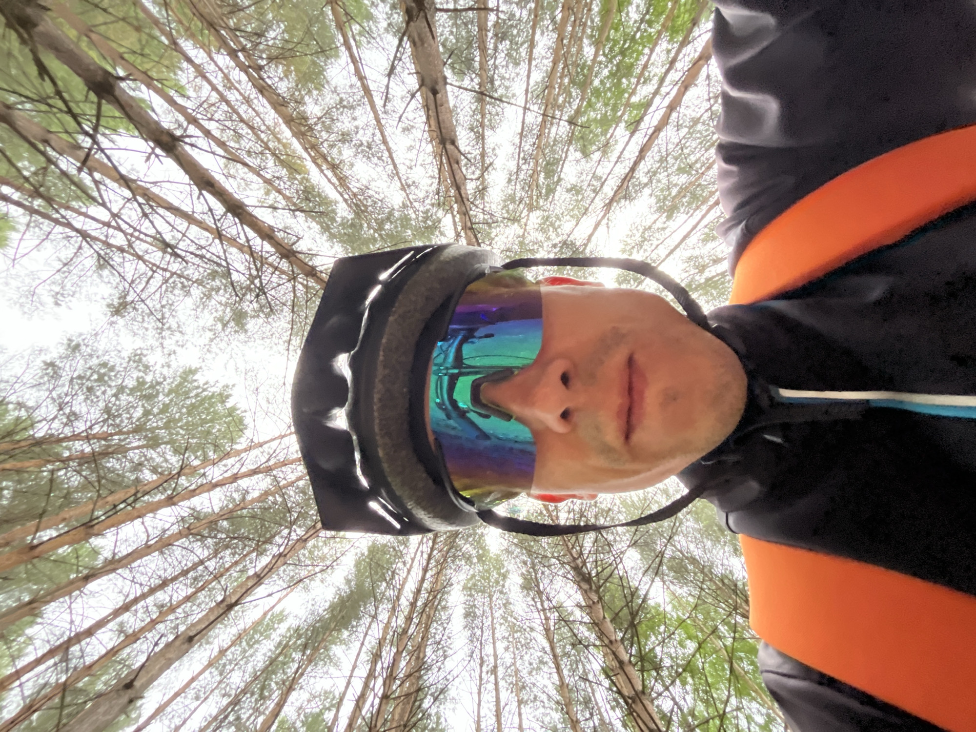 The image is a selfie taken from a low angle, looking up at a person wearing a black helmet and mirrored cycling glasses with a blue and green tint. The person is also wearing a black jacket with an orange strap across the chest. The background consists of the canopy of a forest with tall, thin trees and a bright, overcast sky. The setting appears to be outdoors in a natural environment, likely a forest. There is no visible indication that this location is within the city of Chaikovsky, Russia.