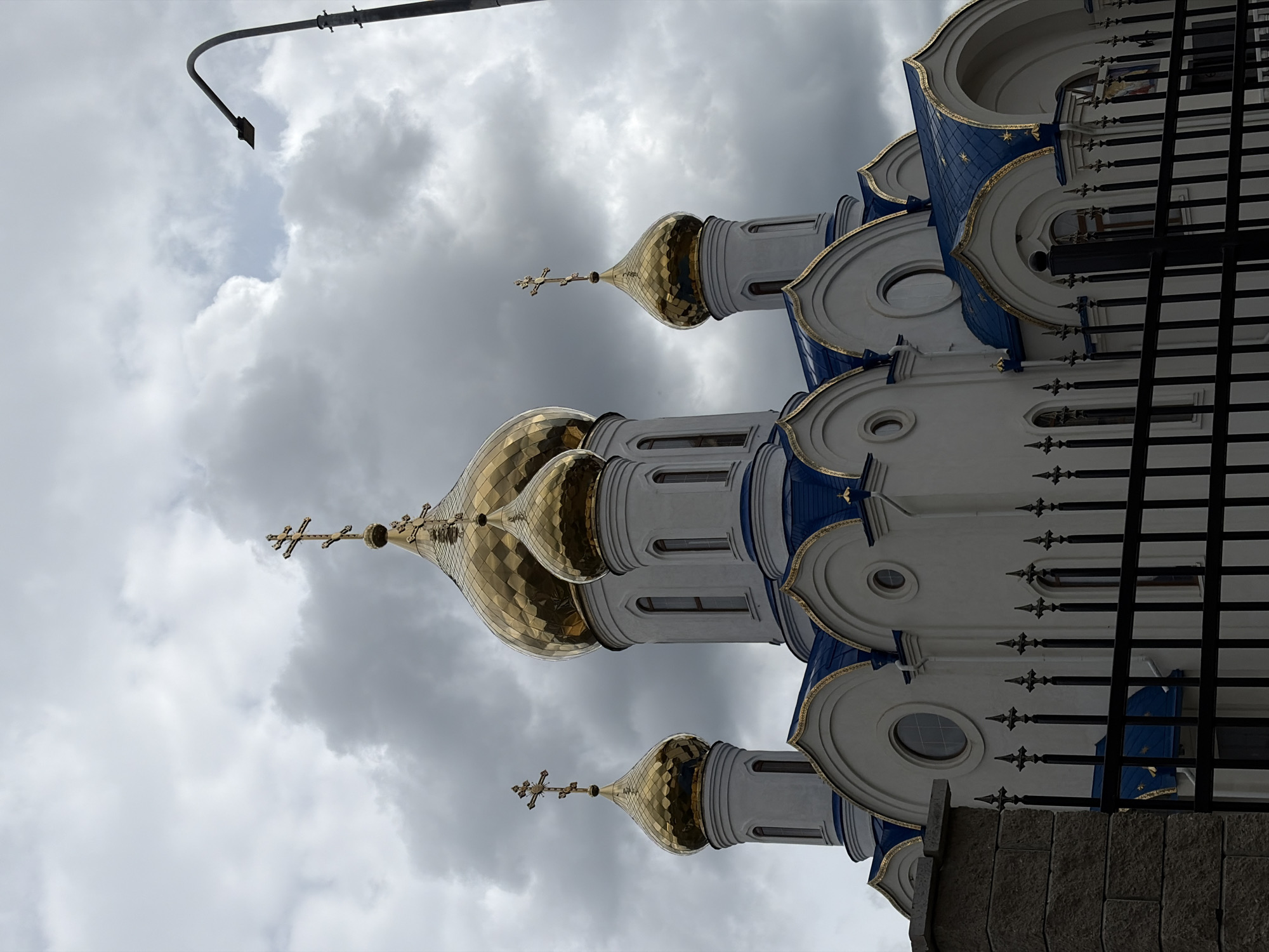 The image is a low-angle shot of the Church of the Nativity of the Blessed Virgin in Novopokrovskaya, Russia. The church features three golden, onion-shaped domes topped with Orthodox crosses. The building is painted in white and blue, with gold trim along the edges. Dark metal fences are in the foreground, partially obscuring the lower portion of the church. Overcast skies are visible in the background, with a street light also appearing in the top left.