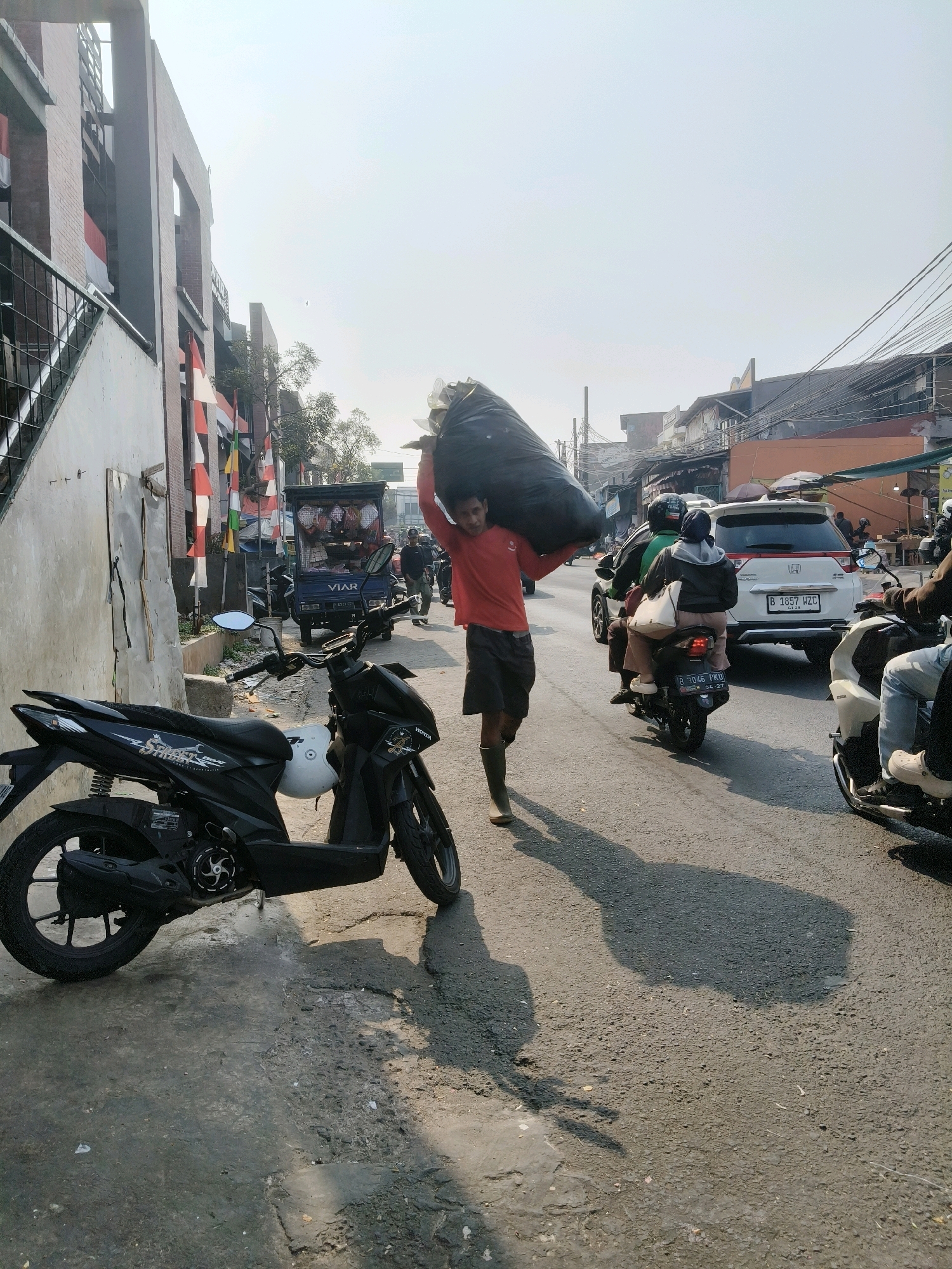 A man in a red shirt and dark shorts carries a large black bag over his shoulder while walking on the side of a street. He is wearing green boots. In the foreground, a black motorcycle is parked on the side of the road. In the background, there is traffic, including cars and motorcycles, and buildings line the street. The scene appears to be in an urban environment in South Tangerang, Indonesia, suggested by the presence of Indonesian flags on some buildings and the style of vehicles and architecture. The sky is bright and hazy, indicating daylight.