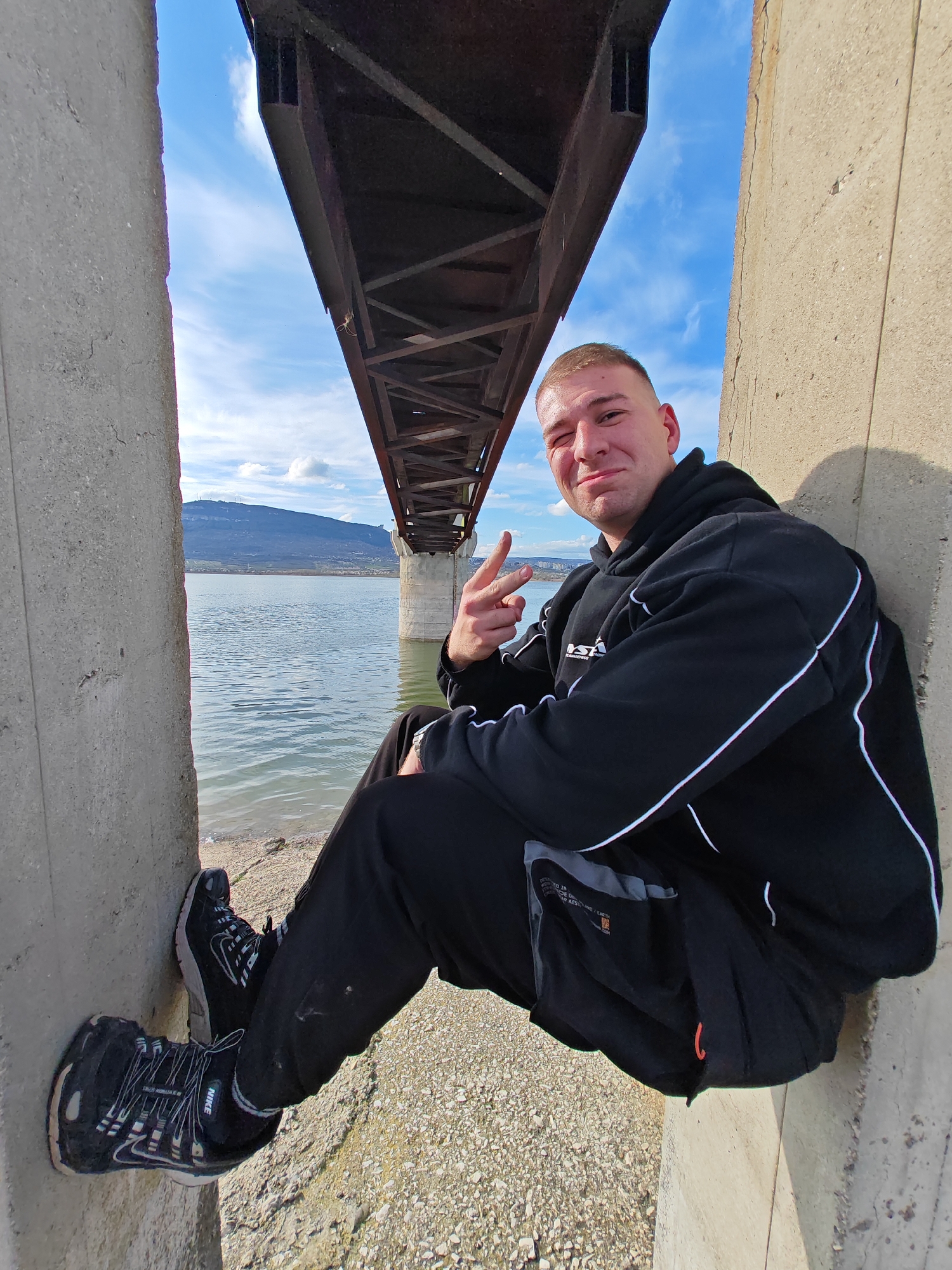 The image depicts a man seated between two concrete pillars under a bridge. The man is wearing a black hooded sweatshirt with white stripes, black pants, and black sneakers with the Nike logo. He is making a peace sign with his left hand and winking. The bridge structure is visible above him, and in the background, there is a body of water, likely a lake or river, with a mountain range in the distance. The ground beneath the man is gravelly. Based on the visible terrain and lake features, it is highly likely this image was taken near the bridge over Yazovir Ticha in the city of Shumen, Bulgaria.