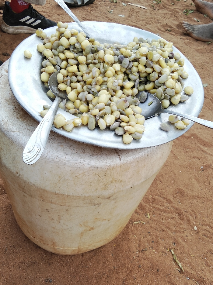 A large, circular metal plate is centrally positioned, filled with a mixture of cooked pale yellow maize kernels and greenish-grey legumes. Two metal spoons are present on the plate; one is partially submerged in the food, and the other rests on the rim, displaying an etched design on its handle. The plate rests on a light-colored, cylindrical plastic container that exhibits surface wear and discoloration. The ground beneath the container is exposed reddish-brown soil, containing minimal scattered debris. In the upper left quadrant, a human foot wearing a black athletic shoe with white stripes and a red sock is visible. In the upper right quadrant, a bare human foot rests on the soil. The scene is located outdoors in the city of Kyamatu, Kenya.
