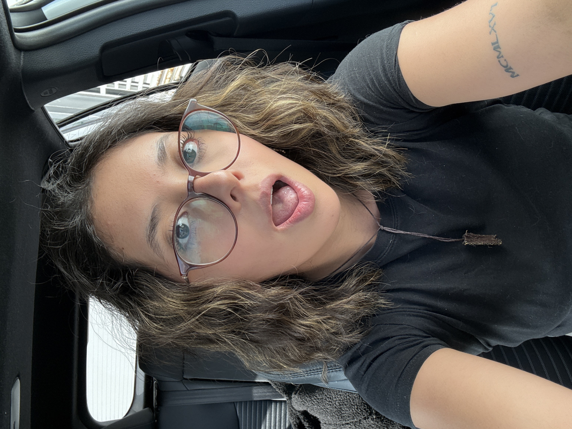 A person wearing glasses and a dark shirt is in a car. The location is Puebla, Mexico. The person has a tattoo on their arm. The person's mouth is open. The car's interior is visible.