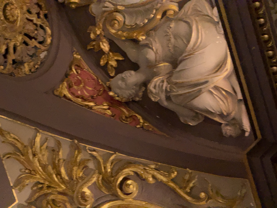 An ornate architectural detail shows classical elements. A sculpted figure, possibly female, drapes in fabric with golden embellishments. Foliage motifs, also in gold, curl around the structure, juxtaposed with a red-painted section. The background is a dark, muted tone, likely part of an interior ceiling or wall. The image captures the grandeur and intricacy of old-world craftsmanship.