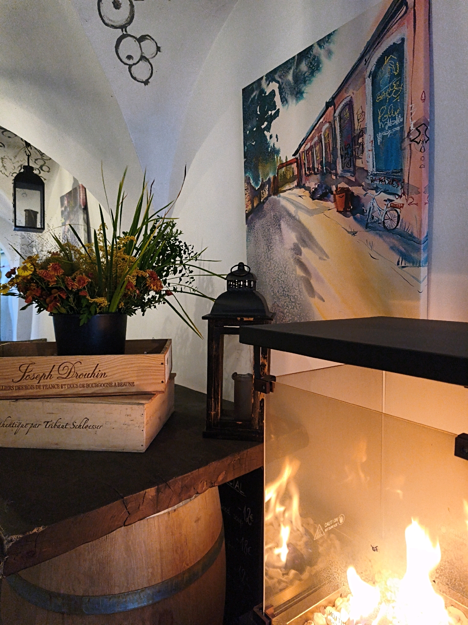 The image depicts an interior scene with a decorative arrangement. In the foreground, a modern fireplace with a glass enclosure radiates a warm glow from its flames. Above the fireplace, a dark countertop supports a wooden crate labeled "Joseph Drouhin" and another crate below it with handwritten text. To the left of the crates, a floral arrangement in a dark pot is placed. Further left, a black lantern hangs from the ceiling. The ceiling itself is arched and painted white, with abstract black drawings of fruit or berries. To the right of the floral arrangement, another black lantern with a glass pane is positioned. In the background, a large, colorful painting of a street scene with a bicycle leaning against a building hangs on the wall. The overall impression is of a cozy and artistically decorated space. The specific location in Tallinn, Estonia, is not discernible from the image itself.