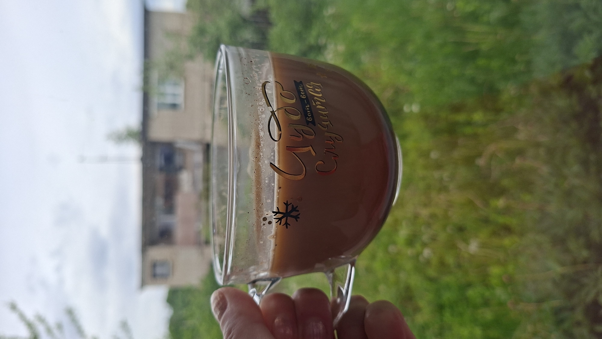 A hand holds a glass mug containing a brown liquid, likely coffee or tea, with a golden inscription in Cyrillic script, including the word "Cozy" and a snowflake symbol, against a blurred backdrop of a residential building and green foliage. The location is Забалоцкі сельскі Савет, Belarus.