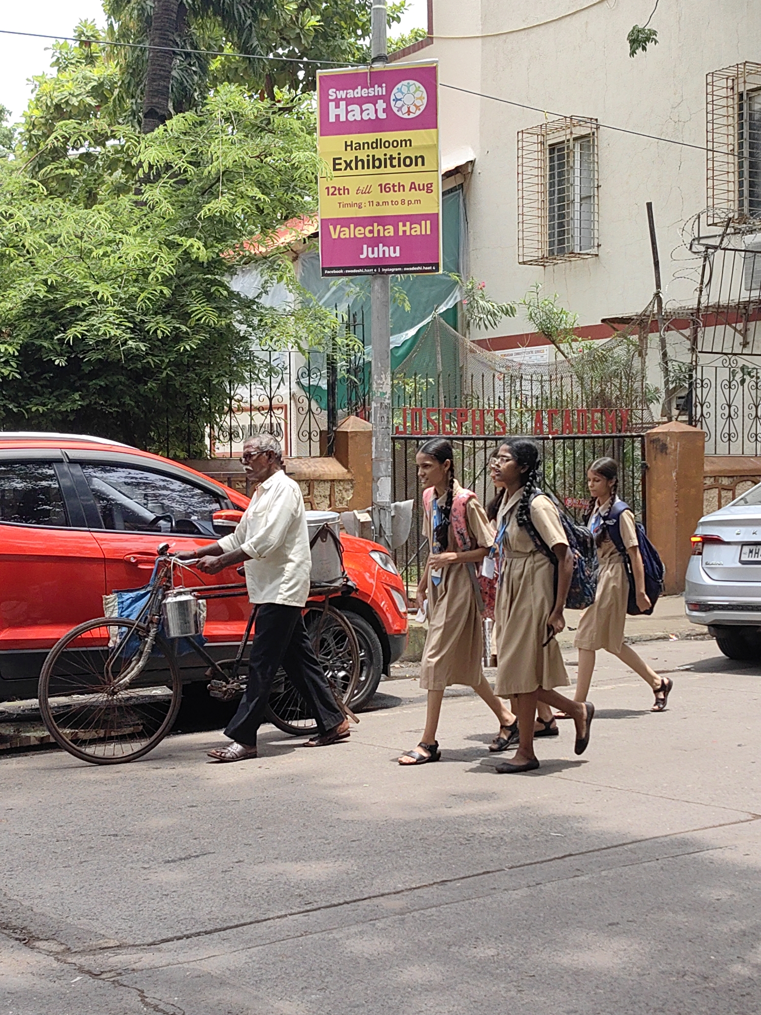 An outdoor, eye-level shot captures a street scene in Mumbai, India, on a bright, hazy day. In the foreground, a man with gray hair and a mustache, wearing a light-colored shirt and dark pants, pushes an old bicycle. The bicycle has a large front wheel, a smaller back wheel, and is laden with two metal containers and a blue bag. To the left of the man and bicycle is the side of a red car.
In the mid-ground, three young girls in school uniforms walk from left to right. They wear light brown dresses, dark socks, and sandals. Each girl has a backpack. The girl in the middle has glasses and her mouth is open as if speaking. Behind the girls is a large metal gate with ornate scrollwork and the words "ST. JOSEPH'S ACADEMY" in red lettering. A street sign that reads "Swadeshi Haat" (Handloom Exhibition, 12th till 16th Aug, Timing: 11 a.m. to 8 p.m., Valecha Hall, Juhu) is visible on a pole to the left. A silver car is parked on the far right.
The background includes a building with barred windows and a tall palm tree to the left. Dense green foliage and smaller trees are also present. The pavement is made of asphalt, and the overall impression is of a busy urban environment.