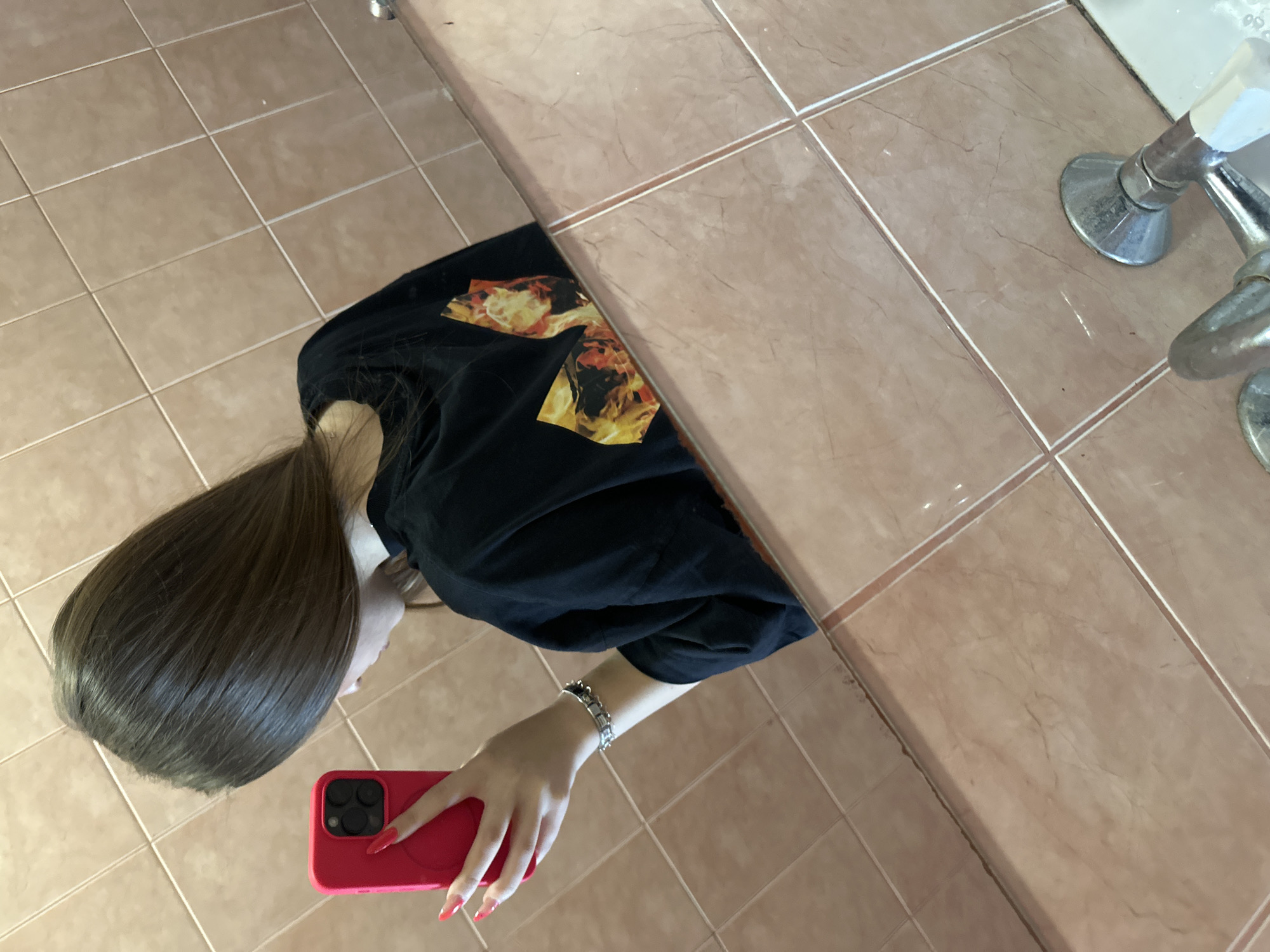 A young woman is taking a selfie in what appears to be a bathroom. The location is possibly in Varna, Bulgaria, but this cannot be definitively determined from the image. The bathroom has light brown, square tiles on the floor and on what appears to be a counter or sink area. A silver faucet fixture is visible. The woman is wearing a black t-shirt with a flame graphic. She has long, straight brown hair. She is holding a red smartphone in her hand, likely using it to capture her reflection. She has a silver bracelet on her wrist and red nail polish. The perspective is from above, looking down at a slight angle. The image appears to be taken using the phone's front-facing camera and is likely a mirror reflection.