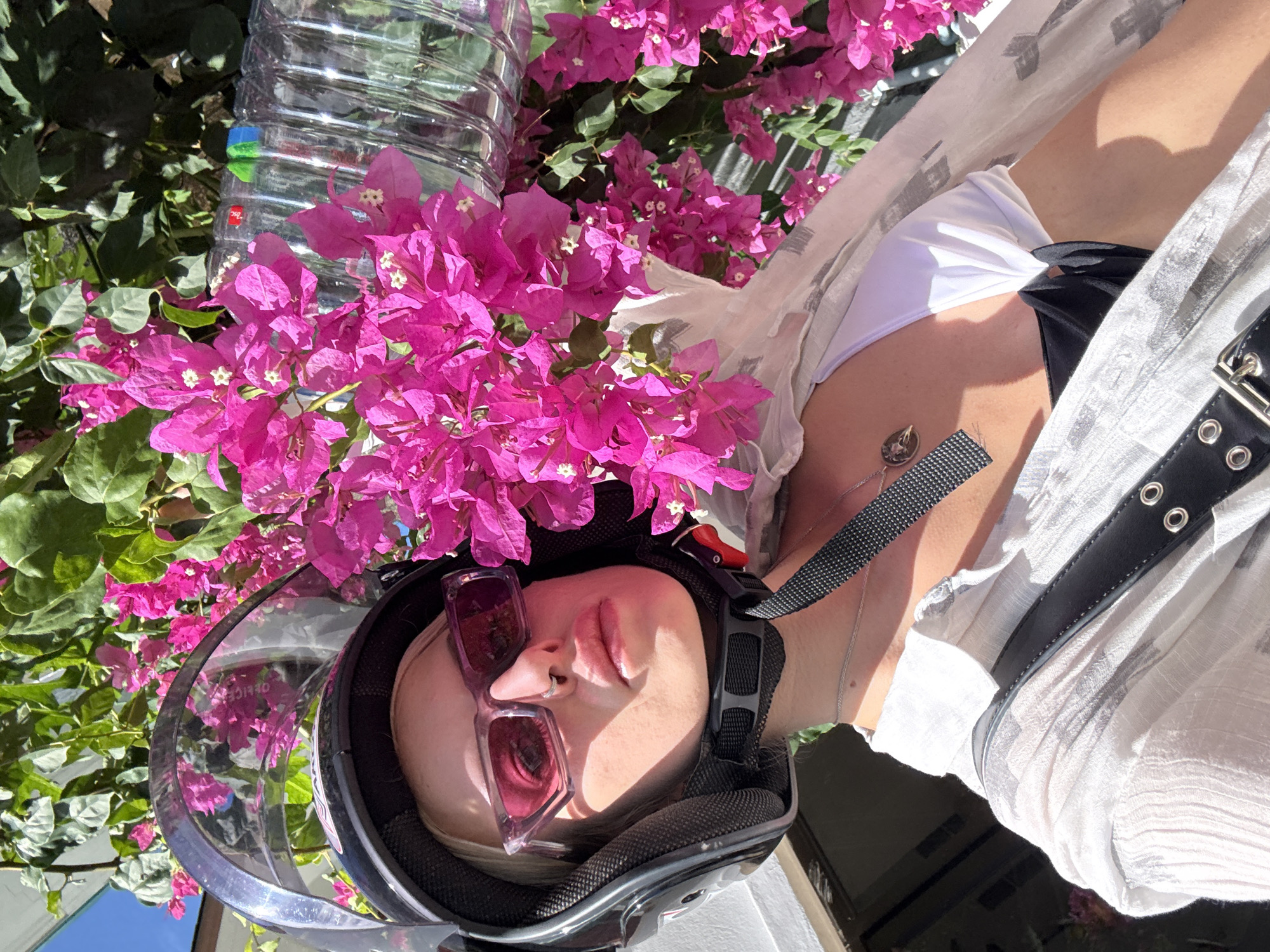 A young woman, wearing a black helmet, pink-tinted sunglasses, and a nose ring, is pictured outdoors in a sunny environment. She is looking directly at the camera with a neutral expression. She is wearing a white bikini top, a light, sheer white shirt, and a black strap with silver hardware across her chest. A silver necklace with a pendant is visible. Behind her, vibrant pink bougainvillea flowers and green leaves create a lush backdrop. A clear plastic bottle with a red and blue cap is partially visible among the flowers. The lighting suggests a bright, warm day. The scene evokes a casual, possibly travel-related context, possibly on the island of Ko Samui, Thailand, given the tropical flora.