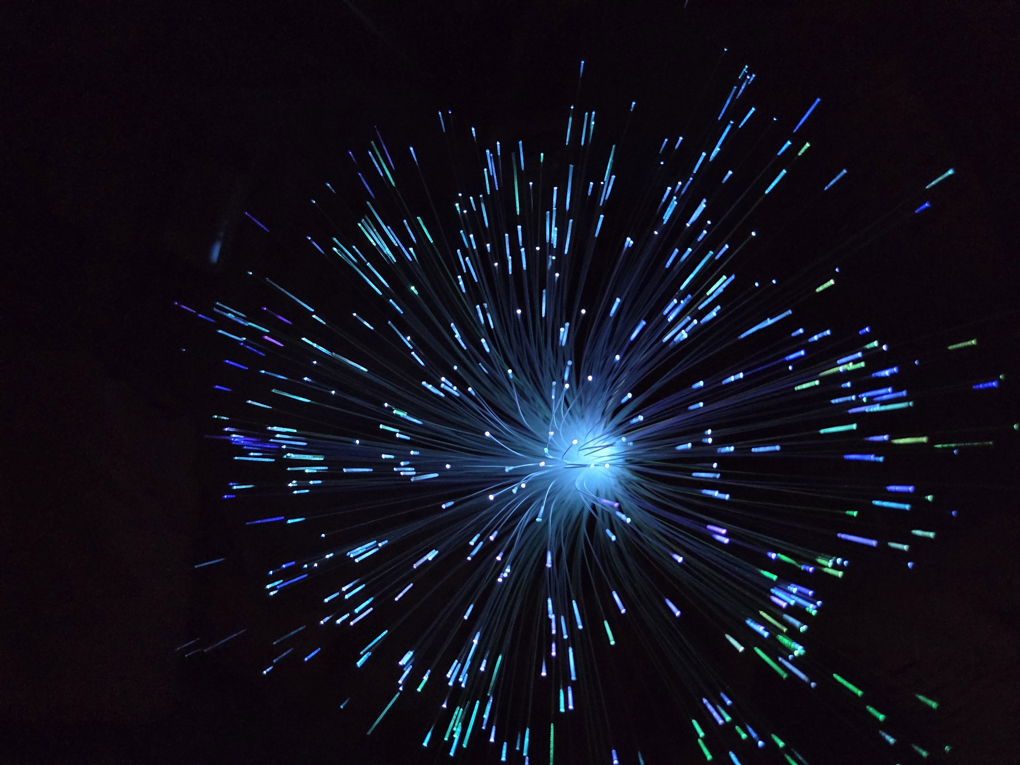 The image depicts a fiber optic light display against a dark background. The light source is centrally located and emits numerous strands of light, predominantly blue, with some strands exhibiting green and purple hues. The light strands radiate outwards from the central source, creating a starburst effect. The background is a dark void, allowing the illuminated strands to stand out prominently. There are no people or other objects visible in the scene. The location of the image is unknown.