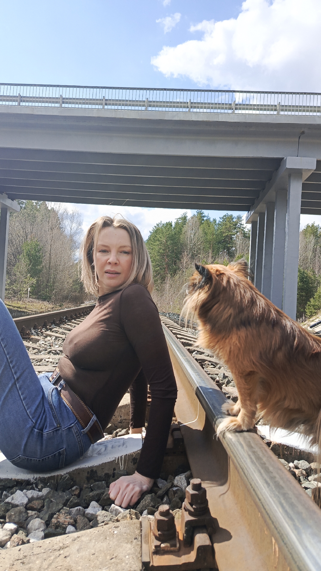 The image features a woman and a dog positioned on railway tracks beneath a concrete bridge in Chaikovsky, Russia. The woman, with blonde hair, is dressed in a dark brown long-sleeved top, blue jeans, and a brown belt. She is seated on the ground beside the railway line, leaning slightly back, and looks directly at the camera with a neutral expression. Her right hand rests on the gravel ballast between the tracks. Beside her, a medium-sized, long-haired dog with reddish-brown fur stands on one of the metal rails, facing away from the camera and looking towards the right side of the frame.
The setting appears to be a natural or semi-rural environment. A dense line of trees, some bare and some evergreen, forms the background behind the railway, suggesting an early spring day. Above the subjects, a large concrete bridge or overpass stretches across the railway, supported by sturdy pillars visible on the right side of the frame. The sky is bright blue with scattered white clouds, indicating clear, sunny weather during the daytime, likely late morning or early afternoon. The railway sleepers, gravel, and metal fastenings are clearly visible in the foreground. There is no discernible text within the image. The scene captures a tranquil, outdoor moment with the woman and her canine companion by the railway.
