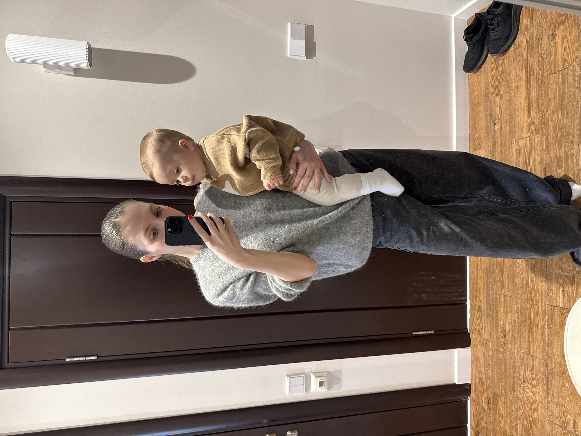 A woman is holding a baby in front of a full-length mirror. The woman is wearing a gray sweater and black jeans. The baby is wearing a tan sweater and white pants. The woman is holding a black smartphone up to take a picture in the mirror. The background shows part of the apartment's interior with a dark brown door and a wooden floor with a pair of black shoes near the wall. The location is likely an apartment somewhere in Russia. There is no definitive evidence of the city.