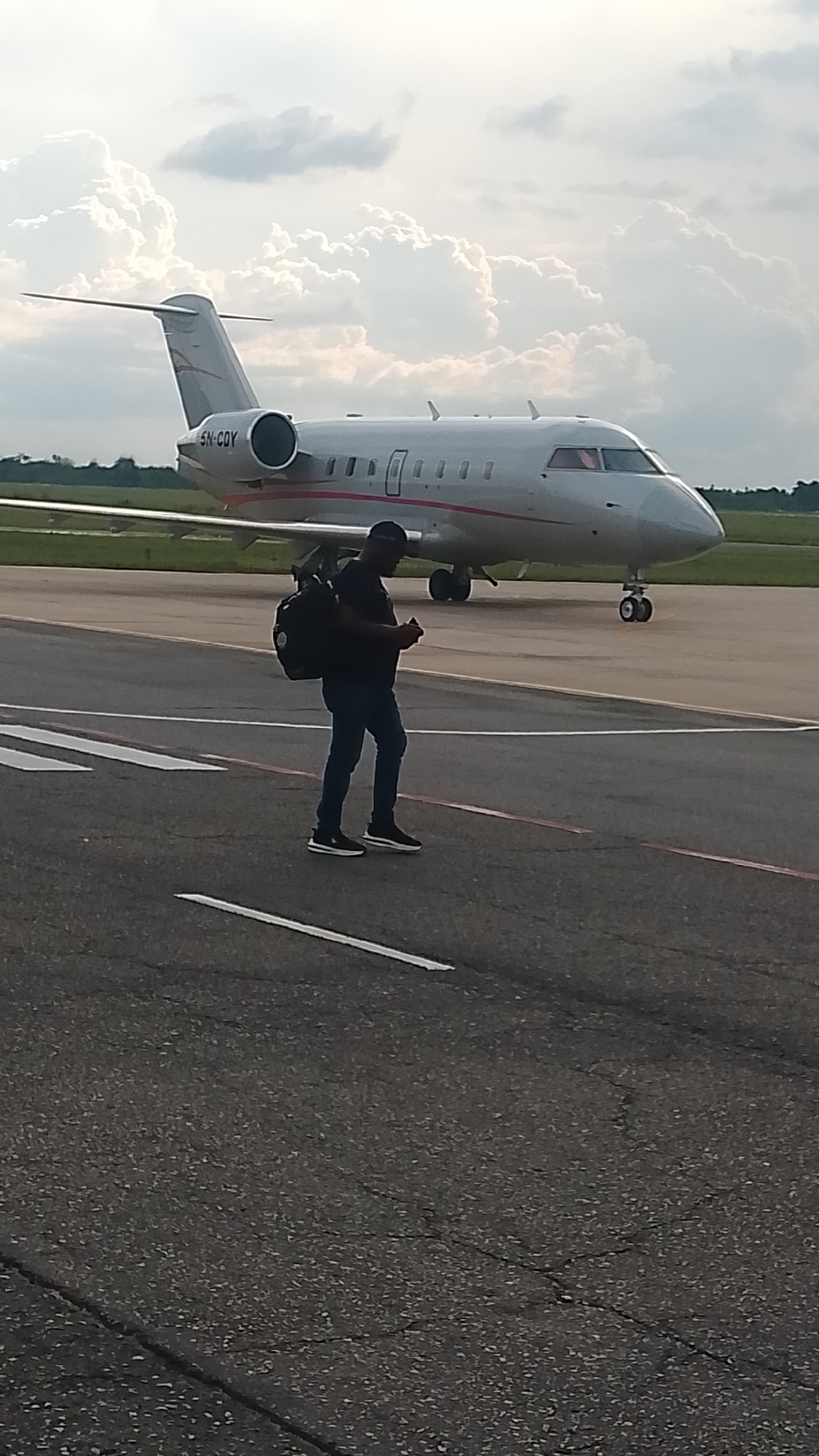 A man walks on a tarmac toward a private jet. The jet is white with a red stripe and the tail number 5N-CQY. The man is wearing dark clothing and carrying a black backpack. The location is likely an airport in or near Orerokpe, Nigeria. The sky is cloudy.