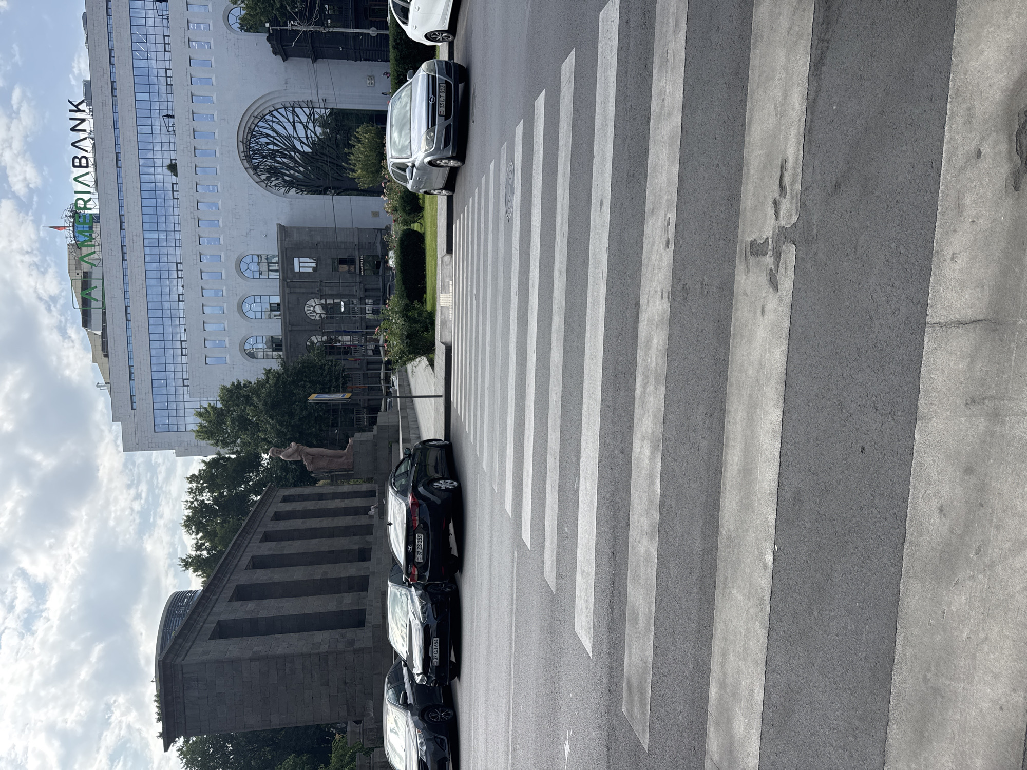 A medium-wide shot captures a pedestrian crossing on a city street in Yerevan, Armenia, with several cars parked on the side of the road. In the background, a large, modern building with the "AMERIABANK" logo is visible, along with a monolithic stone structure featuring several columns. The sky is overcast with scattered clouds. The street appears to be an intersection, and the overall scene suggests a typical urban environment.