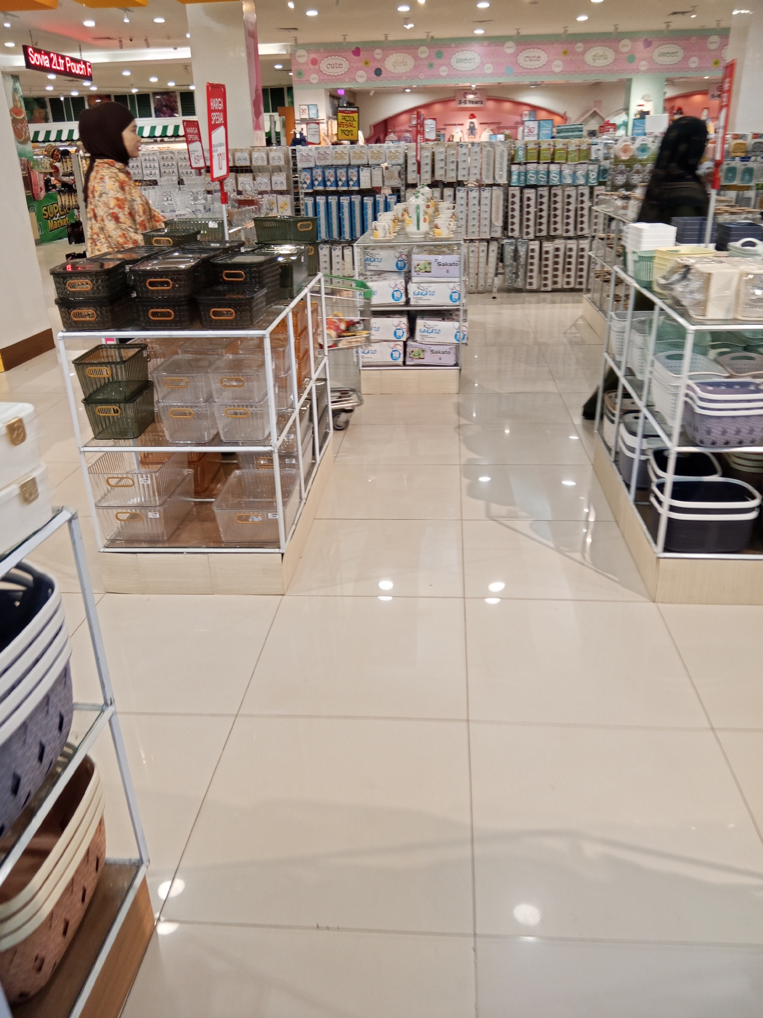 The image shows the interior of a well-lit supermarket or department store, likely a section dedicated to home organization or kitchenware. The flooring is tiled with large, light-colored square tiles that reflect the overhead lighting. In the foreground, a white shelving unit displays various storage containers. Some are dark green, ribbed plastic bins with orange handles, stacked on the top shelf. Below these are clear plastic containers of different sizes, some with orange handles. Further down the shelving unit are more clear plastic storage containers, some with ribbed detailing.
To the left of this shelving unit, a portion of another shelving unit is visible, holding stacked white containers with gold clasps. Also on the left, a person wearing a hijab and a patterned orange and cream top is visible, appearing to browse or shop. They are facing away from the camera and towards the displays.
Further into the store, more shelving units are visible, displaying a variety of products, including what appear to be packaged household goods and kitchen items. Some displays have colorful signage. In the background, more shelves with boxed items are visible, and a glimpse of another person in a dark outfit can be seen.
The overall impression is of a clean, organized retail environment with a wide selection of products. The lighting is bright and even, creating a slightly reflective surface on the floor. The presence of storage containers suggests a focus on organization and household goods.
Based on the signage visible, specifically the "Sovia 2Ltr Pouch" sign, and the general appearance of the store, it is highly probable that this location is a department store or a large supermarket in Lubuklinggau, Indonesia, such as a branch of Transmart, Hypermart, or a similar retail establishment.