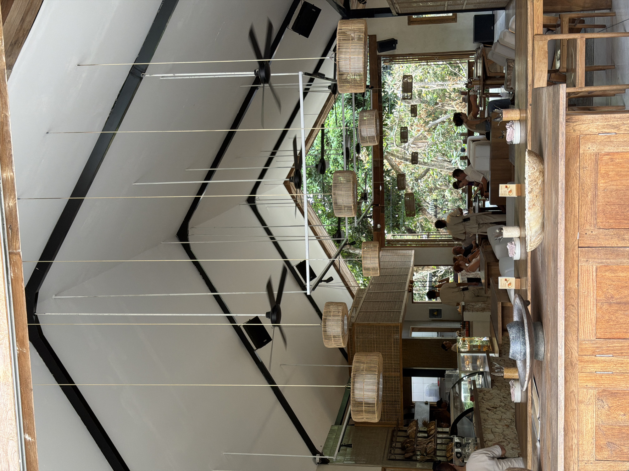 The image depicts the interior of a restaurant with a high, vaulted ceiling, framed by exposed black beams. Multiple pendant lights with rattan shades hang from the ceiling, along with two ceiling fans. A large window at the back of the restaurant offers a view of lush green foliage, suggesting an outdoor or garden setting.
Inside, several patrons are seated at tables, engaged in conversation or dining. A person in a light-colored uniform appears to be serving or attending to customers. In the foreground, a wooden counter or table is visible, adorned with decorative items. Behind the counter, a display case with food items and a coffee machine are present, indicating a food service area. The overall ambiance appears relaxed and inviting, possibly a cafe or a restaurant in Kuta Selatan, Indonesia, given the tropical vegetation visible through the window.
