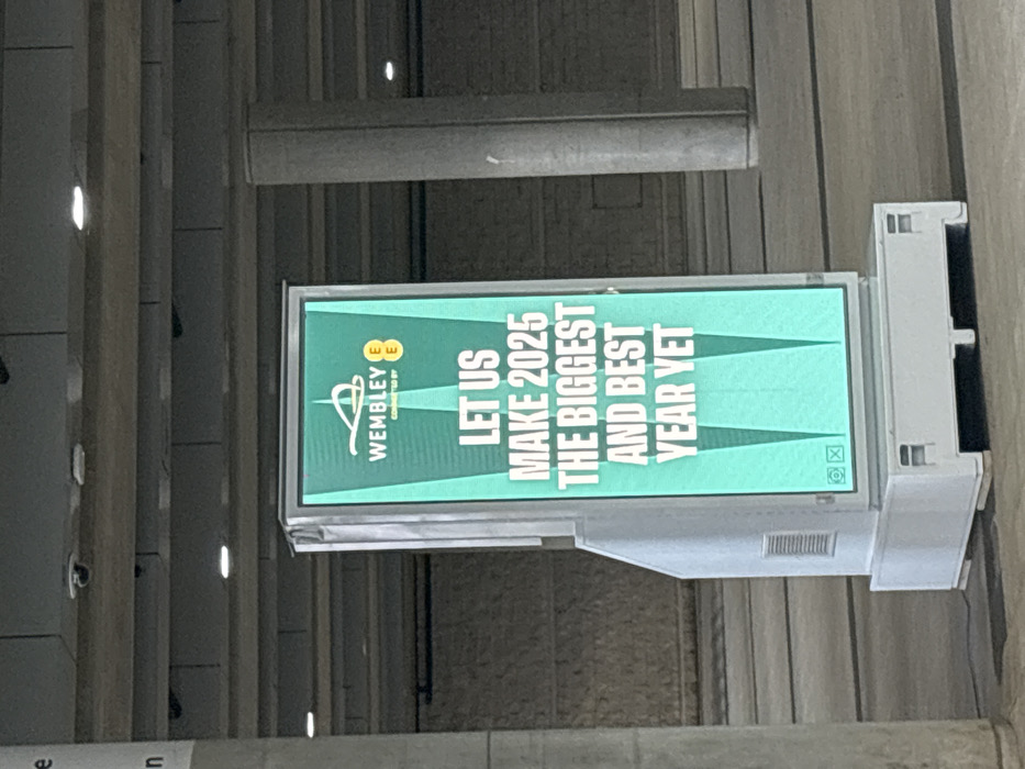 An indoor scene displays a brightly lit digital advertisement board mounted vertically on a structural element, possibly a wall or pillar, within a public concourse or building. The background shows neutral-colored architectural features, including horizontal beams and textured walls, typical of a large venue. No people are visible.
The screen exhibits a teal green background with white text and logos. At the top, the "Wembley" logo is prominently displayed, followed by "CONNECTED BY EE," with the EE logo also visible. The main message on the board reads: "LET US MAKE 2025 THE BIGGEST AND BEST YEAR YET." The text is large and sans-serif, arranged vertically, indicating an aspirational message for the upcoming year. Below the text, two green triangular shapes point upwards. In the bottom right corner of the screen, faint small icons are visible, likely generic display controls or status indicators. The environment appears well-lit by artificial lighting. Given the "Wembley" branding, the location is confirmed to be at Wembley in the United Kingdom, likely within Wembley Stadium or an associated complex.