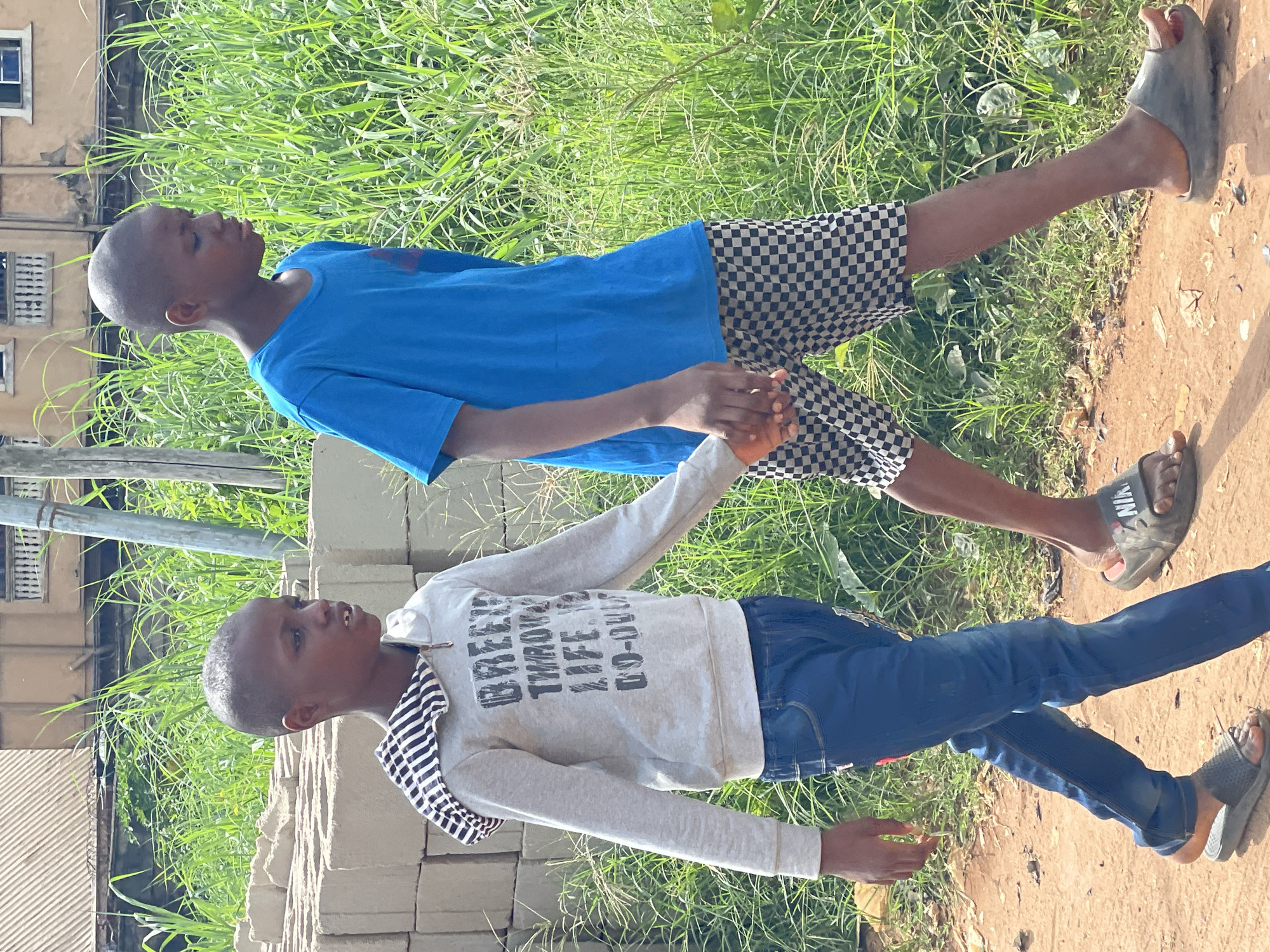 Two young boys are holding hands. One boy is wearing a blue shirt and checkered shorts. The other boy is wearing a grey sweatshirt with the phrase "BREEZY THROUGH LIFE CO-ORD" and blue jeans. Both are wearing sandals. They stand on a dirt path, with lush green grass on one side and a building under construction with concrete blocks on the other. The image was taken in Nkpor, Nigeria.
