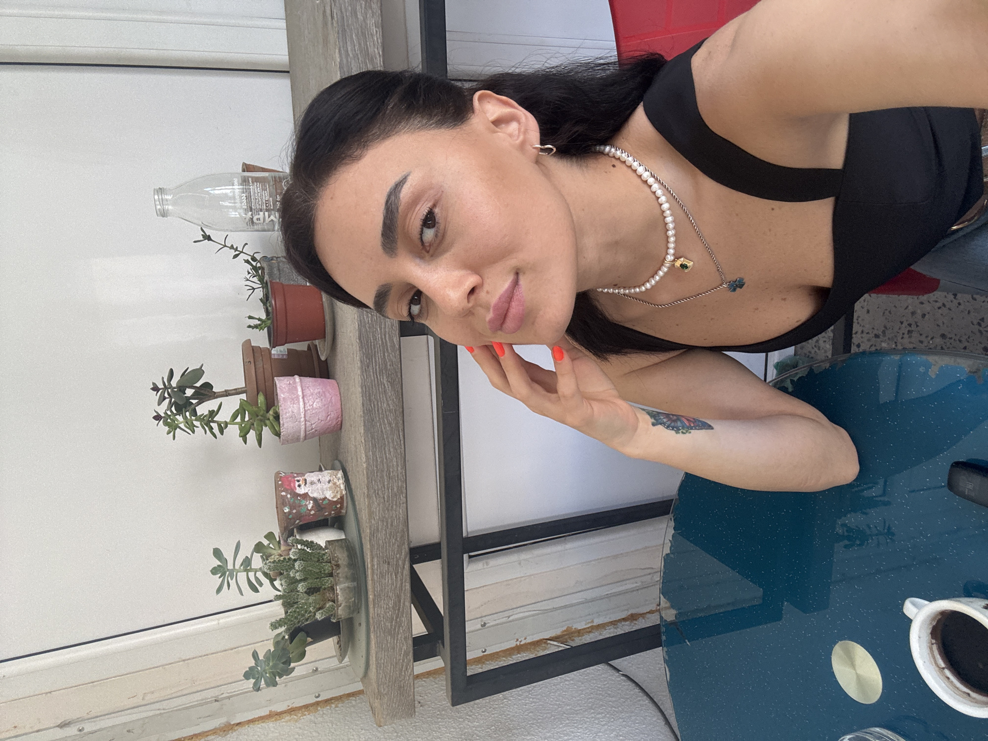 A woman is taking a selfie at a table with a dark blue surface. She has long, dark hair, dark eyes, and is wearing a black top and jewelry. Her left hand is near her face, and her right arm rests on the table. On the table, there is a white cup with a dark liquid, likely coffee, and a black object. Behind her, a shelf holds several potted plants, including succulents and a small cactus. A clear glass bottle is also on the shelf. The background appears to be an outdoor setting with a white wall and a door. The scene is lit by natural light. No specific details about the city of Tbilisi, Georgia, are visible in the image.