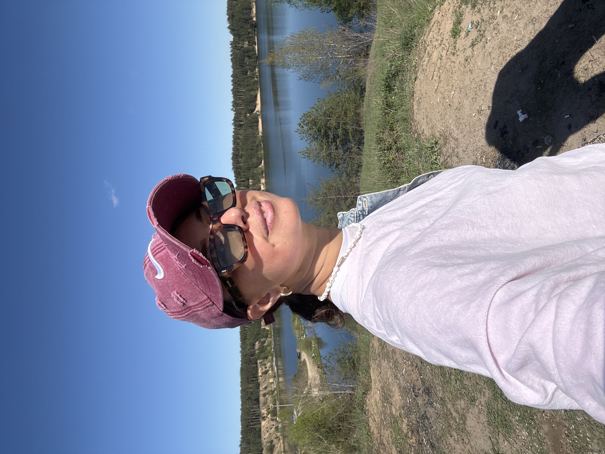 A woman is standing outside on a sunny day. She is wearing sunglasses and a pink baseball cap, with a white shirt and pearl necklace. In the background is a body of water with trees lining the far shore. The location is outside the city of Kazan, Russia. The ground is dirt and grass. The woman’s shadow can be seen on the ground.