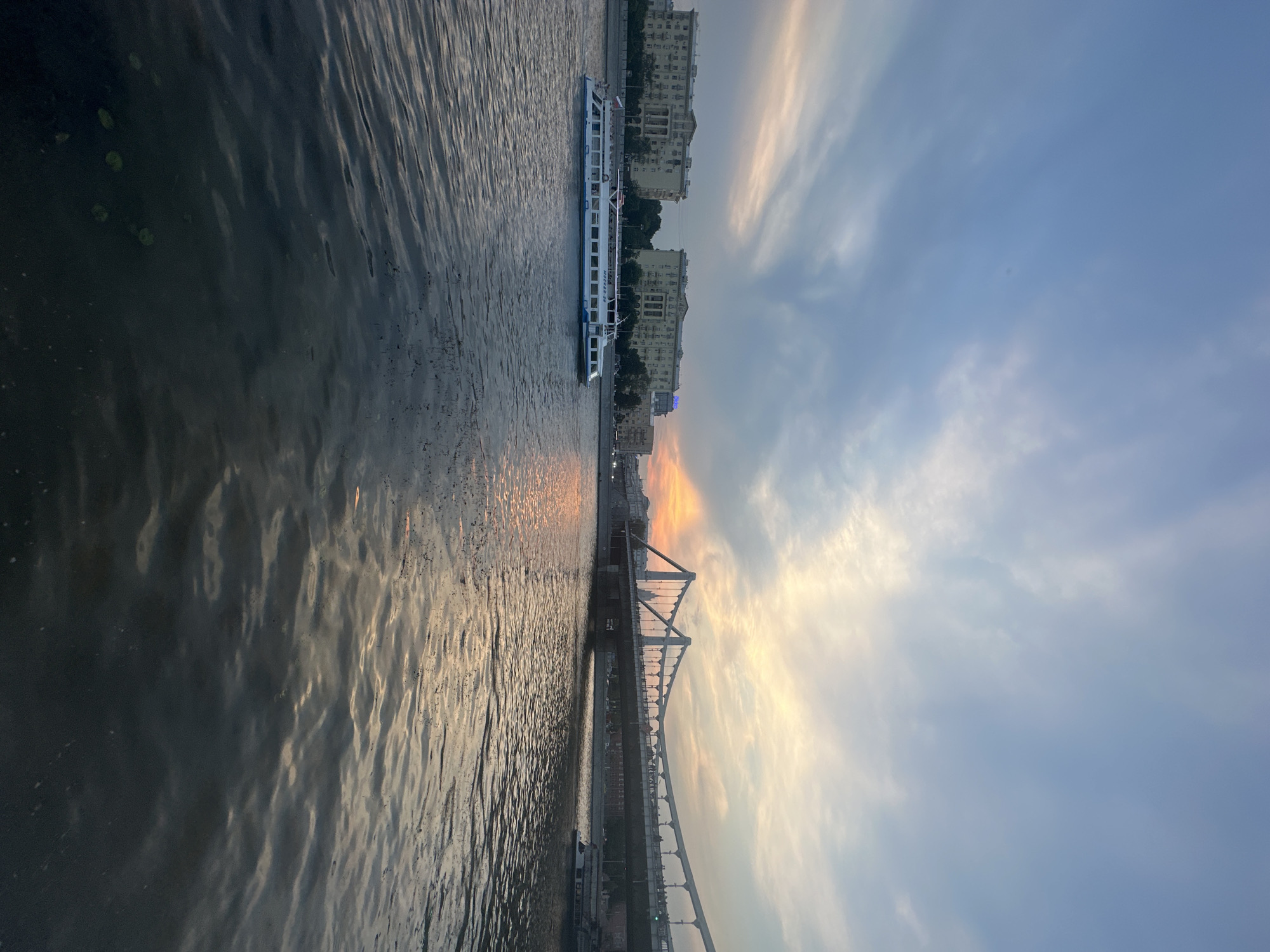 This vertical, eye-level shot captures a wide view of a river at dusk. A white passenger boat with blue trim is moving from left to right in the foreground, on the water. Across the river, on the embankment, are several large, light-colored buildings with many windows. To the right of the buildings, a long suspension bridge spans the river, with people visible walking across it. The sky above is a mix of blue and gray clouds, with a warm orange glow from the setting sun peeking through the clouds on the horizon, casting reflections on the water. The water itself is dark and rippled, with some debris floating on the surface in the foreground. The scene appears to be urban, likely a riverfront in a city, but the specific location is unknown.