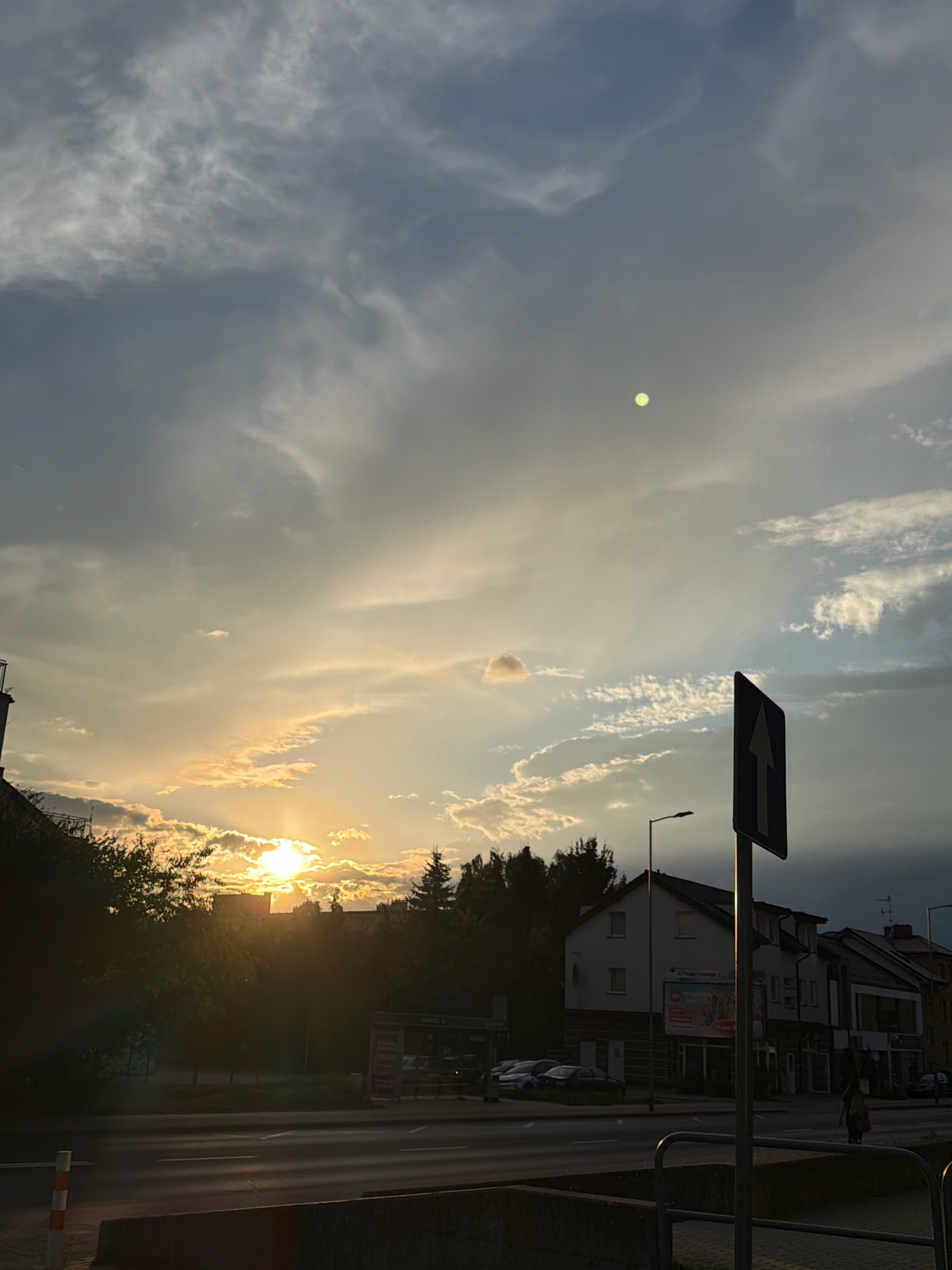 The image depicts a street scene in Koszalin, Poland, during sunset. The sky is filled with a mix of grey and white clouds, with the sun breaking through on the left side, casting a warm glow. A faint, circular object, possibly the moon or a lens flare, is visible in the upper right quadrant of the sky.
In the foreground, a street is visible with lanes marked by white lines. Cars are parked on the side of the road, and buildings line the street, some appearing to be shops with advertisements. A traffic sign with an upward-pointing arrow is prominently displayed on a pole. Trees and greenery are present, particularly on the left side, silhouetted against the setting sun. A lamppost stands near the buildings. A person is walking on the sidewalk on the right side of the image. The overall atmosphere is that of a typical urban environment at dusk.