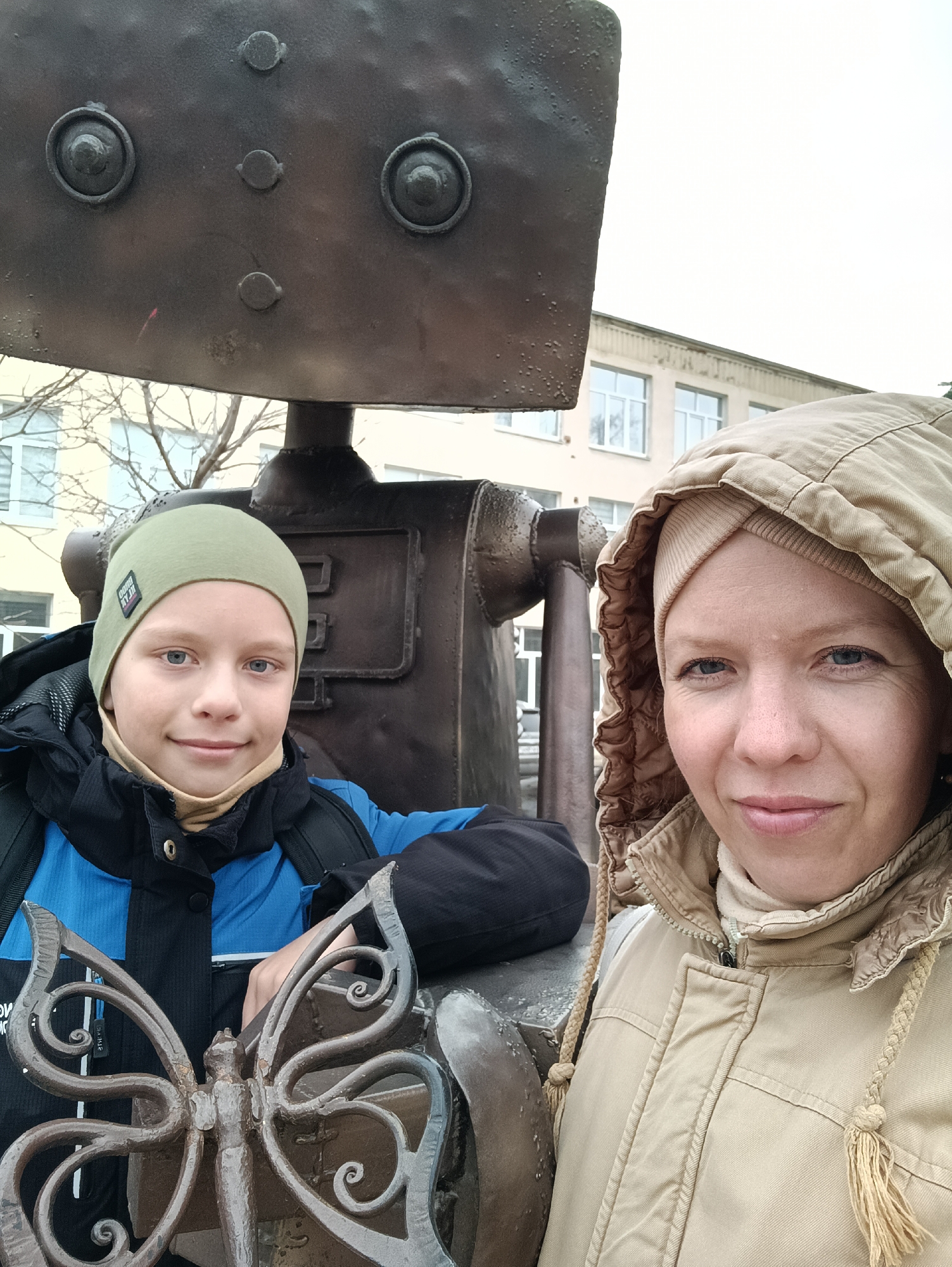 This outdoor selfie captures a woman and a boy in Chaikovsky, Russia, posing beside a large, dark metal sculpture. The sculpture, possibly depicting a robot or industrial figure, features prominent bolted elements resembling eyes and other facial characteristics.
On the right, a woman with fair skin and blue eyes looks at the camera with a gentle smile. She is wearing a beige hooded jacket with braided drawstrings and a beige beanie or headband under her hood.
To her left, a boy with fair skin and blue eyes is also looking at the camera with a pleasant smile. He is dressed in a green beanie, a blue and black jacket, and a light brown neck gaiter. On his green beanie, the text "GOOD VIBES" is visible. His hand rests on an intricately designed, dark wrought iron butterfly ornament located in the foreground.
The setting appears to be a cool or cold day, indicated by the heavy outerwear worn by both subjects and the bare tree branches visible in the background. An overcast sky suggests daytime. Behind them, a light-colored building with multiple windows, possibly a school or public institution, stands under the muted sky. The overall scene conveys a calm and positive mood.