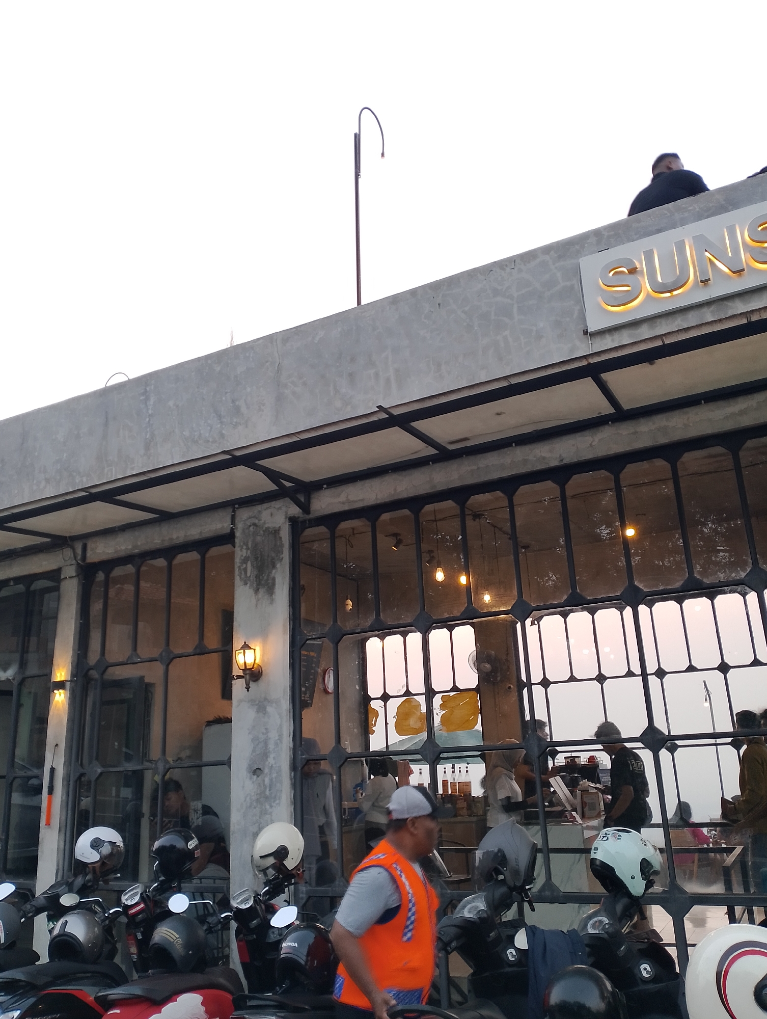 The image depicts the exterior of a building, likely a cafe or restaurant, in Srimulyo, Indonesia. The building features a concrete facade with large, industrial-style windows framed by black metalwork. The name "SUNS" is prominently displayed on a lit sign above the entrance. In the foreground, several motorcycles are parked, some with helmets on their seats. People are visible inside the building, some behind a counter, suggesting service is being provided. The lighting and atmosphere suggest it is late afternoon or early evening. A street lamp is visible on the roof of the building.