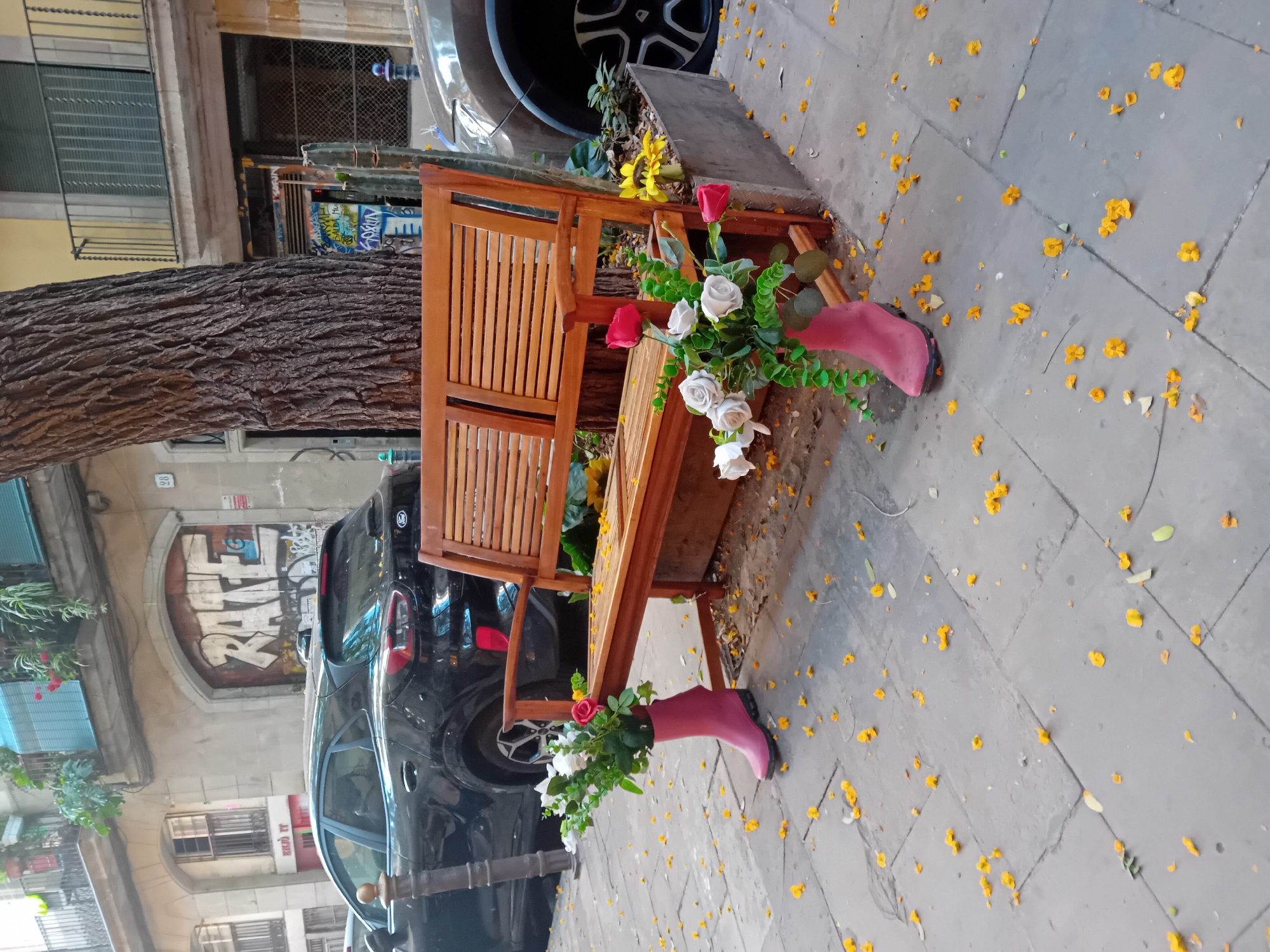 This vertical image depicts a decorative urban street scene in Barcelona, centered around a whimsical display on a sidewalk. The ground is paved with large, light-grey flagstones, extensively scattered with bright yellow flower petals, suggesting a nearby flowering tree or recent wind.
The main subject is a light-brown wooden bench, designed with a planter box integrated into its structure, filled with yellow flowers and green foliage. On either side of the bench, two pink rubber boots (wellies) are creatively repurposed as planters, each holding a bouquet of artificial flowers including white and red roses mixed with green leaves.
To the left of the bench, a large tree trunk with thick, textured bark is prominent. Behind the bench and tree, several dark-colored cars are parked along the street. The background features typical urban architecture with multi-story buildings, some displaying balconies and window grates. On a building wall behind one of the cars, graffiti is visible on a closed door or archway, with the word "RAFAEL" clearly discernible in large white letters. A small number "20" is also visible on an adjacent building. The scene is illuminated by soft, natural light, indicating daytime.