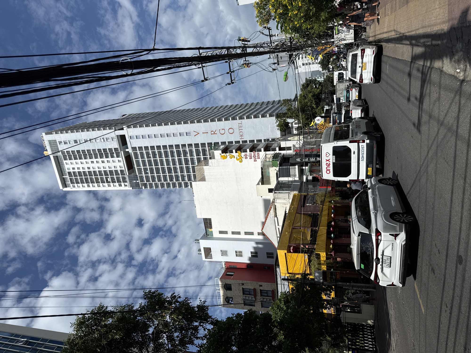 A street scene in Nha Trang, Vietnam, is captured under a bright, partly cloudy sky. The primary focus is a tall, modern white hotel building with the "VIRGO HOTEL" logo vertically inscribed on its facade. Surrounding the hotel are other buildings of varying heights and colors, including a striking yellow structure and a red building. The street is paved and appears to be a busy thoroughfare, with several vehicles present. In the foreground, a white SUV is parked on the right side of the road, and a white van with "anex" branding is driving away from the camera. Other cars and vehicles are visible further down the street. Electrical wires are strung overhead, creating a dense network above the scene. Pedestrians are present on the sidewalk to the right, with one woman in black shorts and a tank top walking away from the camera, and another child to her right. A man is walking on the sidewalk on the left side of the street, partially obscured by trees. The environment suggests an urban setting with a mix of commercial and residential structures.