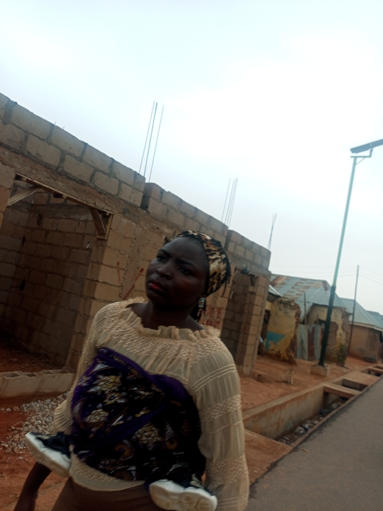 A woman wearing a headscarf and a light-colored blouse is standing in what appears to be a residential area in Tempure, Nigeria. She is holding a patterned cloth and shoes. Behind her is a partially constructed brick building, and across the street are more buildings with corrugated metal roofs. A streetlight is visible in the background, and there is a narrow concrete drainage ditch running alongside the street.