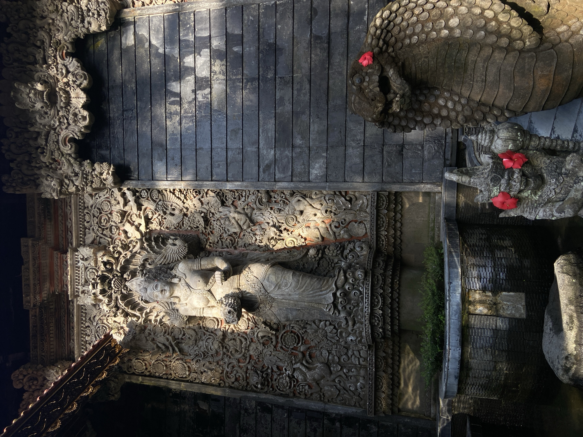 This image captures an ornate, open-air setting in what appears to be a traditional Indonesian building, possibly a temple or a decorative courtyard. The shot is taken from above.
The composition features a variety of textures and materials, including elaborately carved stone or concrete structures, dark wooden planks, and greenery. At the lower-left side is a detailed bas-relief depicting a reclining figure. Adjacent to the planks is a stone serpent. The scene includes vibrant red flowers. The water feature includes some greenery.
Based on the architectural style and the presence of traditional carvings and water features, the location is likely in the Ubud District, Indonesia, known for its rich cultural heritage and traditional Balinese architecture.