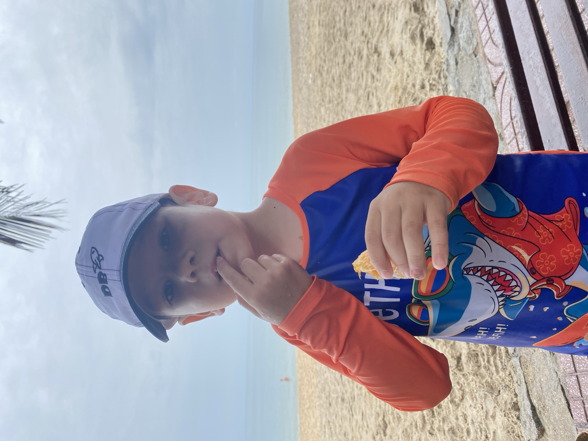 A young boy in Phu Thuy Ward, Vietnam, sits on a bench at a beach. He wears a blue and orange long-sleeved rash guard with a cartoon shark design and a gray baseball cap with the letters "DB" on it. The boy is eating an orange, holding a piece in his hand and touching his lips with his fingers. The beach is sandy, and the sky is overcast. In the distance is the sea.