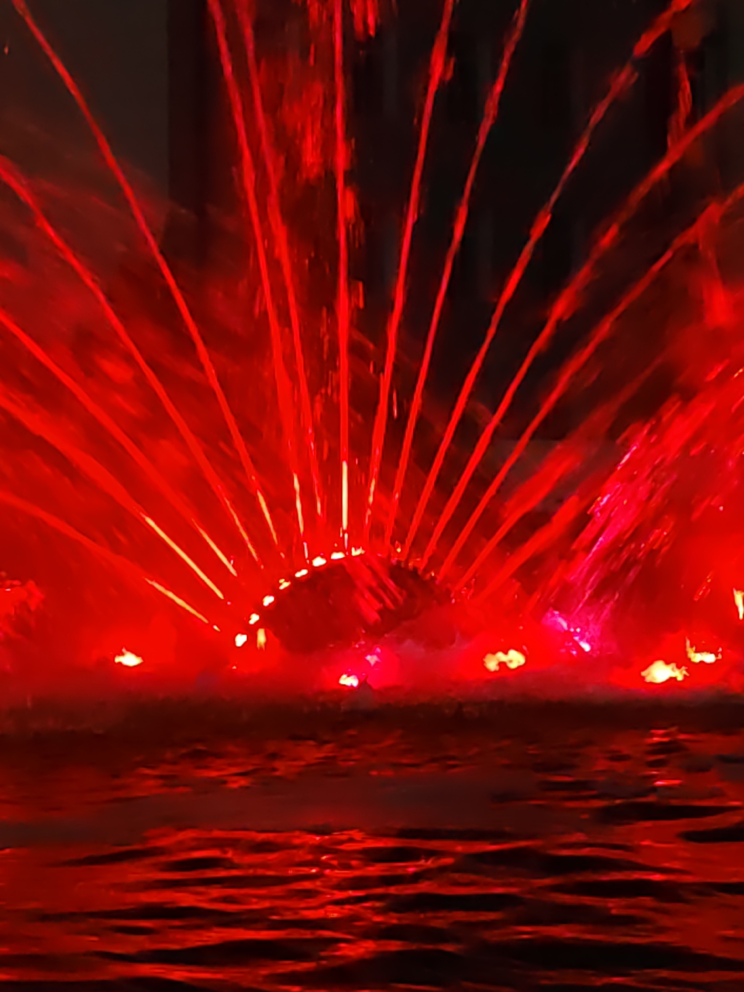 The image depicts a close-up, vertical shot of a fountain at night, illuminated by red lights. Water streams are spraying upwards and outwards, creating a fan-like effect. The water droplets and streams are highly reflective, catching the red light and creating a vibrant, almost laser-like appearance. The background is dark, suggesting a nighttime setting. The water surface in the foreground is rippling, also reflecting the red light, giving it a textured, luminous quality. There are no people or identifiable landmarks visible in the image. Based on the provided information, this scene is located in Rostov-on-Don, Russia.