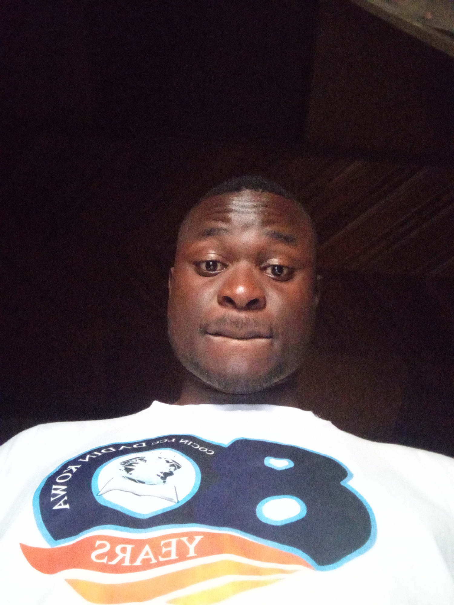 A close-up, head-and-shoulders selfie taken from a low angle shows a Black man from the chest up. He has a shaved head and is looking directly at the camera. He is wearing a white t-shirt with a graphic design on the front. The graphic includes the text "8 YEARS" in large, orange, yellow, and white letters, with a circular emblem above it. The emblem is dark blue with white trim and contains a black and white illustration of a face. To the left of the emblem, in white, is the text "AWOK Y", and to the right, also in white, is "COCIA LEE DVDIH KOMA" in reverse. The man's skin tone is dark, and he has a short mustache. The background is dark and appears to be an indoor setting, possibly a room with wooden walls. The lighting is dim, with the man's face illuminated.