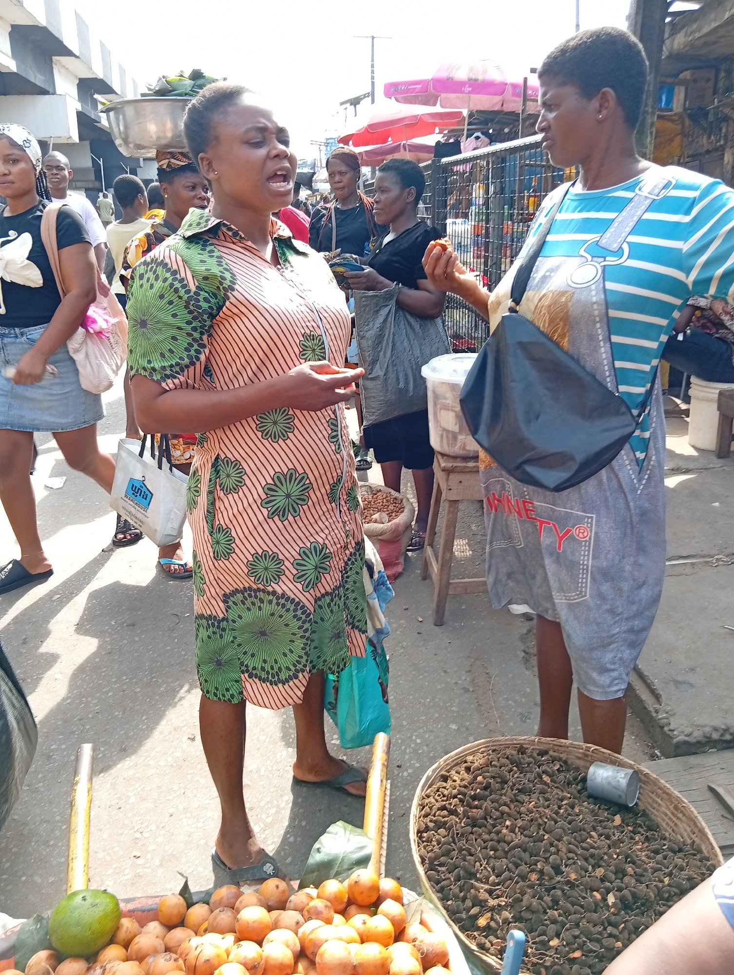 The image depicts a street scene in Nkpor, Nigeria. Two women are the primary focus. One woman, wearing a peach and green floral print dress, is speaking with animation, gesturing with her hand. The other woman, wearing a blue and white striped top and a gray denim-style apron with the word "NINETY" printed on it, is listening to the other woman. A black bag hangs over her shoulder. In the foreground, a cart holds a pile of orange fruits and a single green fruit and a basket filled with dark brown seeds. Other people are visible in the background, including women carrying bags and objects on their heads. The background shows a metal fence, vendor stalls with pink umbrellas, and the partial view of a building, suggesting a busy market area. The setting is outdoors with bright sunlight illuminating the scene.