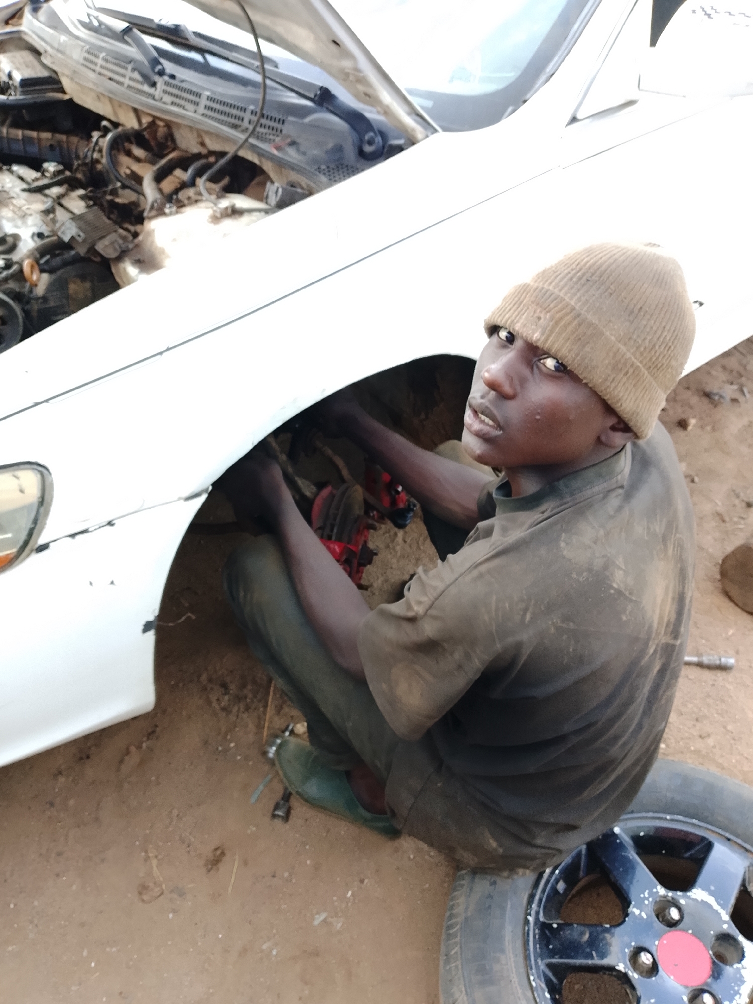 A young male wearing a brown knitted cap and a dark, dirt-stained t-shirt and pants is crouched down in front of a white car with its hood open. He appears to be working on the car's brake assembly, which is visible and painted red. The ground is dirt, and a spare tire is lying on the ground next to him. The scene is likely outdoors in a workshop or repair area.