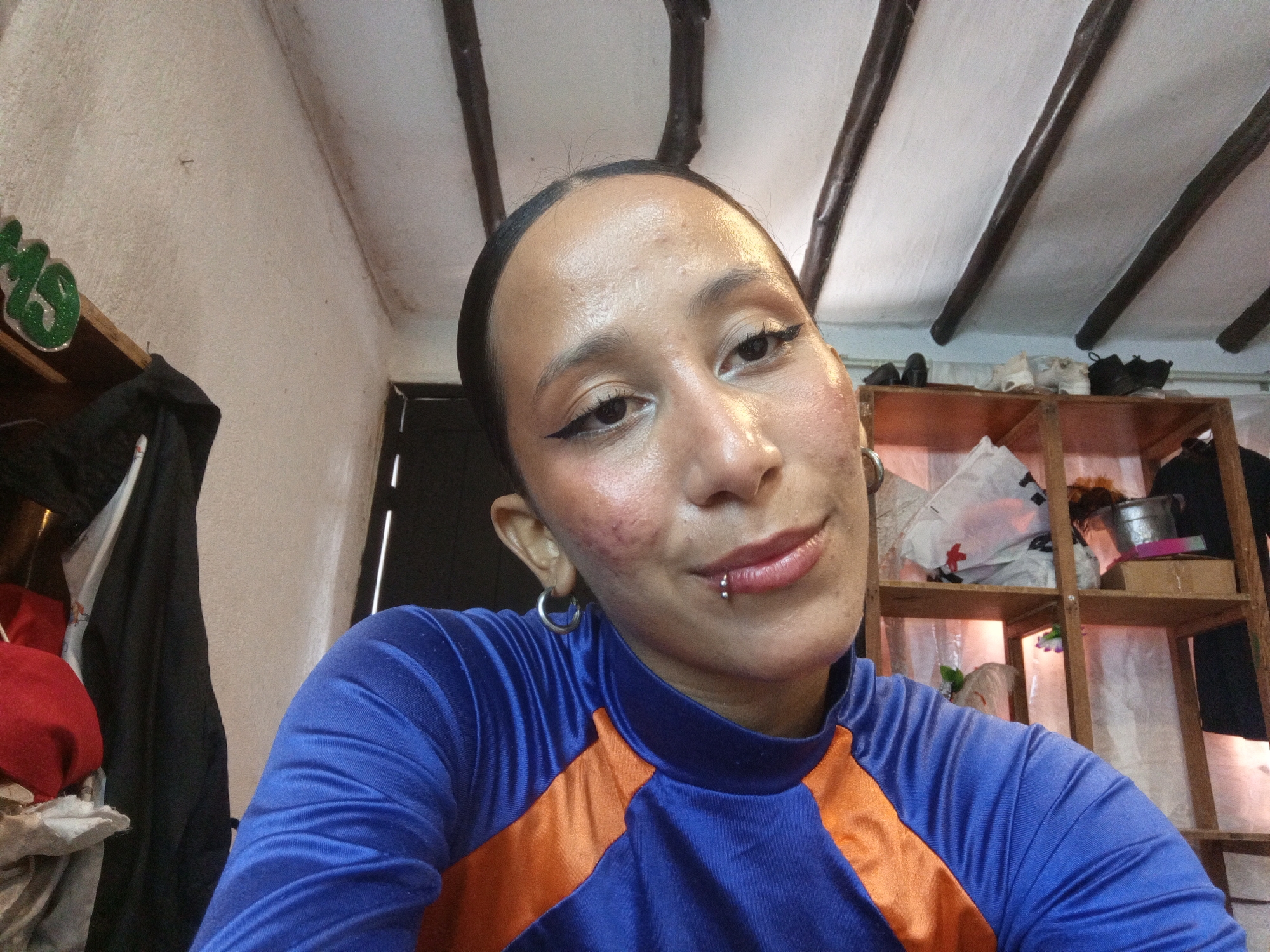 A close-up selfie of a person with tan skin wearing a blue and orange shirt. The person has dark hair slicked back, a small hoop earring, and a lip piercing. They are facing the camera with a slight smile. The background includes a white wall, a wooden ceiling with dark beams, and a wooden shelf containing various objects. The location is potentially Porlamar, Venezuela, but there is no evidence of the location within the photo itself.