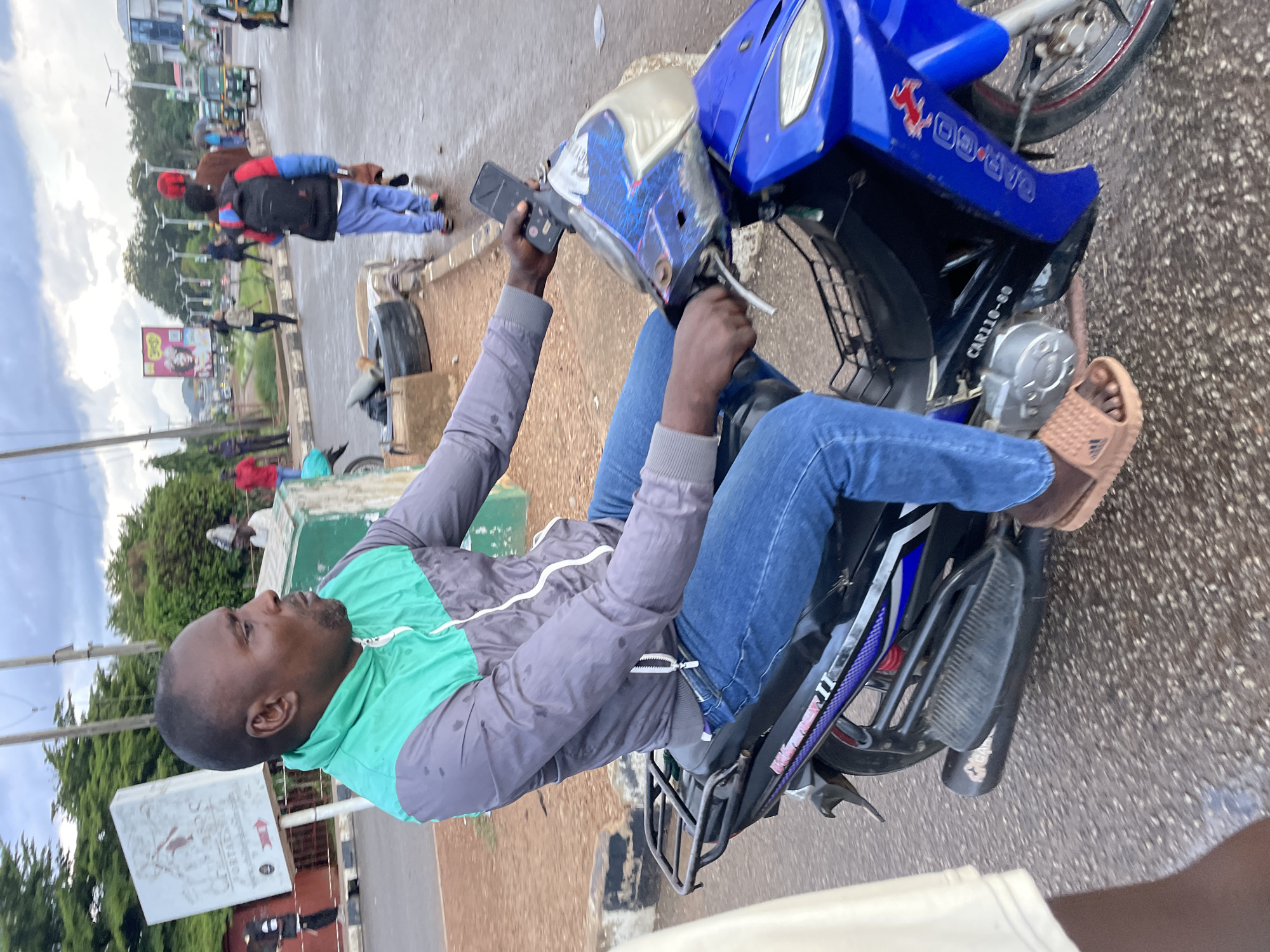 The image shows a man riding a blue motorcycle in Jos, Nigeria. He is wearing a grey and green jacket and blue jeans. The man is holding a black smartphone in his right hand. The motorcycle has a black rear rack and a blue fairing. In the background, there are other vehicles and pedestrians. A signpost with "PORTALS" is visible on the left. The ground is wet, suggesting recent rain.