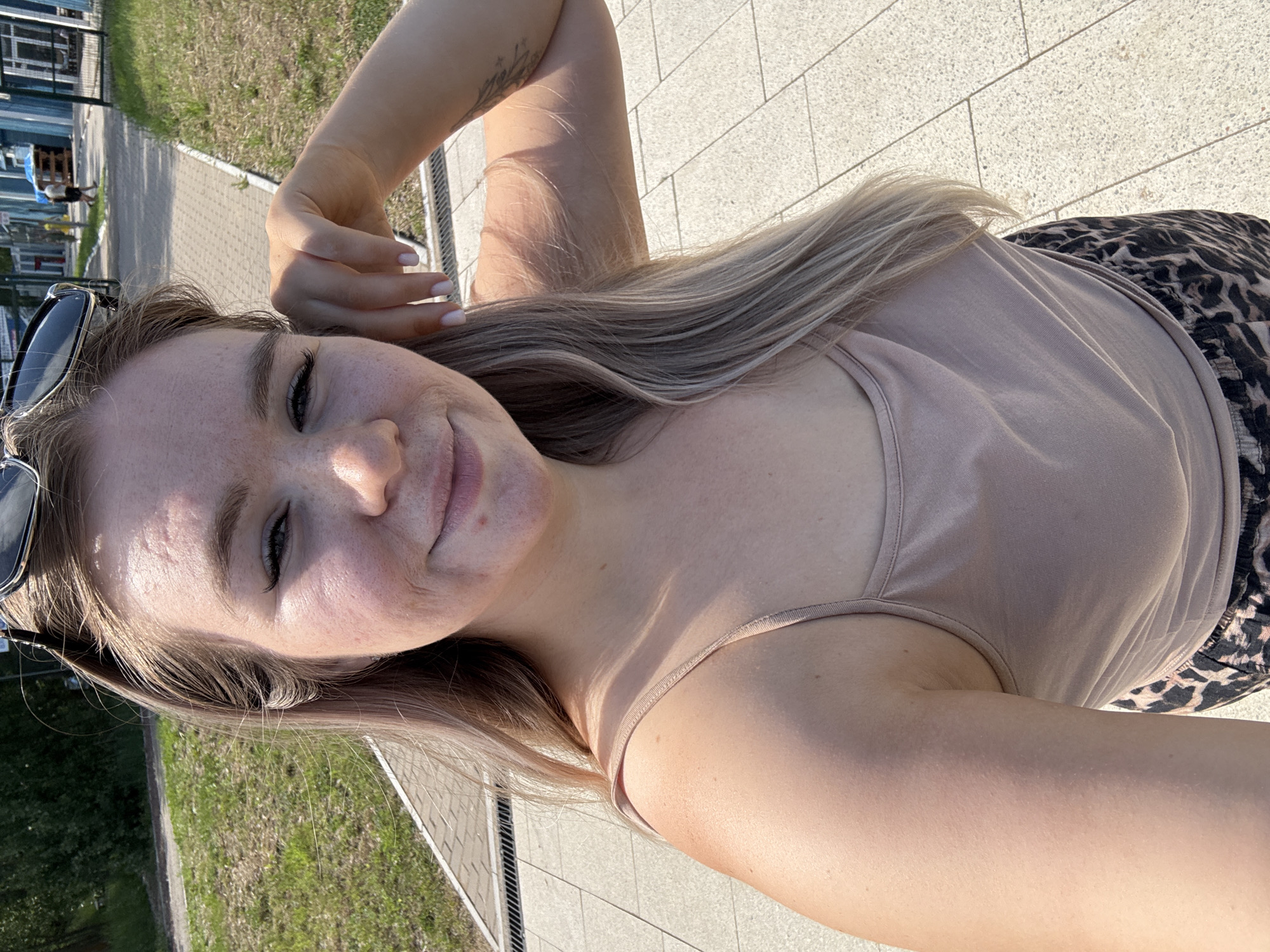 A young Caucasian woman with blonde hair and freckles is taking a selfie outdoors on a sunny day. She is wearing a light brown tank top and leopard-print shorts. Her left hand is resting on her head, and her right arm is bent at the elbow. Sunglasses are perched on her head. She appears to be standing on a paved walkway with a grassy area to her left and buildings in the background. The location is likely near a commercial area, possibly a park or public space, in Chaikovsky, Russia, given the context.