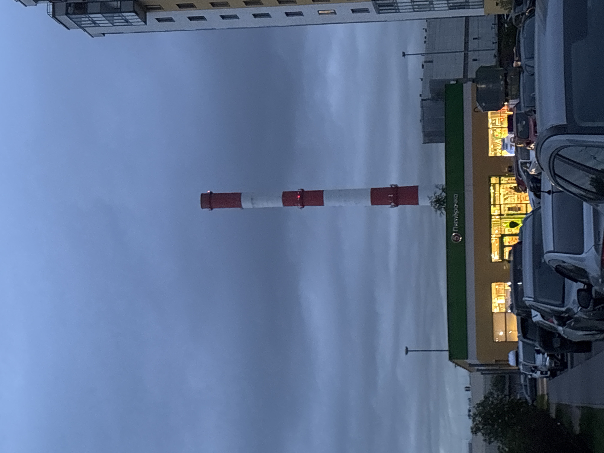 The image shows a cityscape, likely in the city of Saint Petersburg, Russia. The sky is overcast with a dominant gray color.
In the foreground, a large, striped red and white industrial chimney stands prominently in the frame. It is tall and cylindrical.
To the right of the chimney, there is a supermarket or commercial building with illuminated windows, suggesting it's dusk or evening. The building has a green facade. Rows of parked cars are visible in front of the building.
In the upper-left corner, a section of an apartment building is visible.