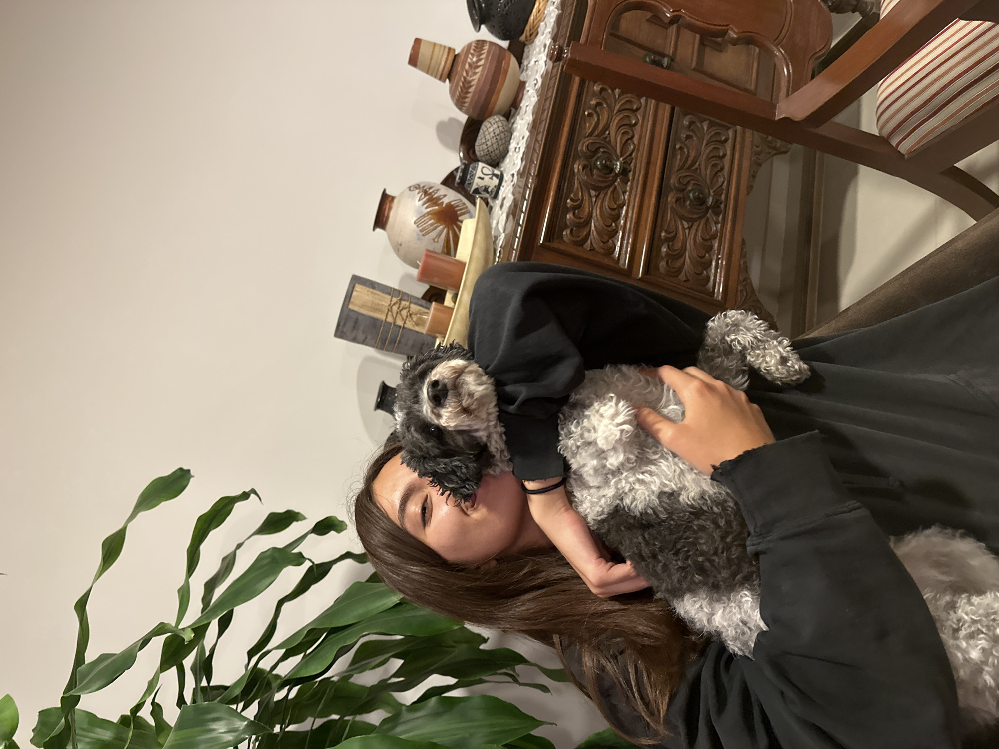 A person with long brown hair is holding a black and white dog in their arms. The person is wearing a black sweatshirt. Behind them, there is a plant with long green leaves, a decorative cabinet, and various vases and decorative items. The location is likely indoors, potentially a home. It is not possible to identify the city of Barrio de Nuevo León, Mexico from the image.