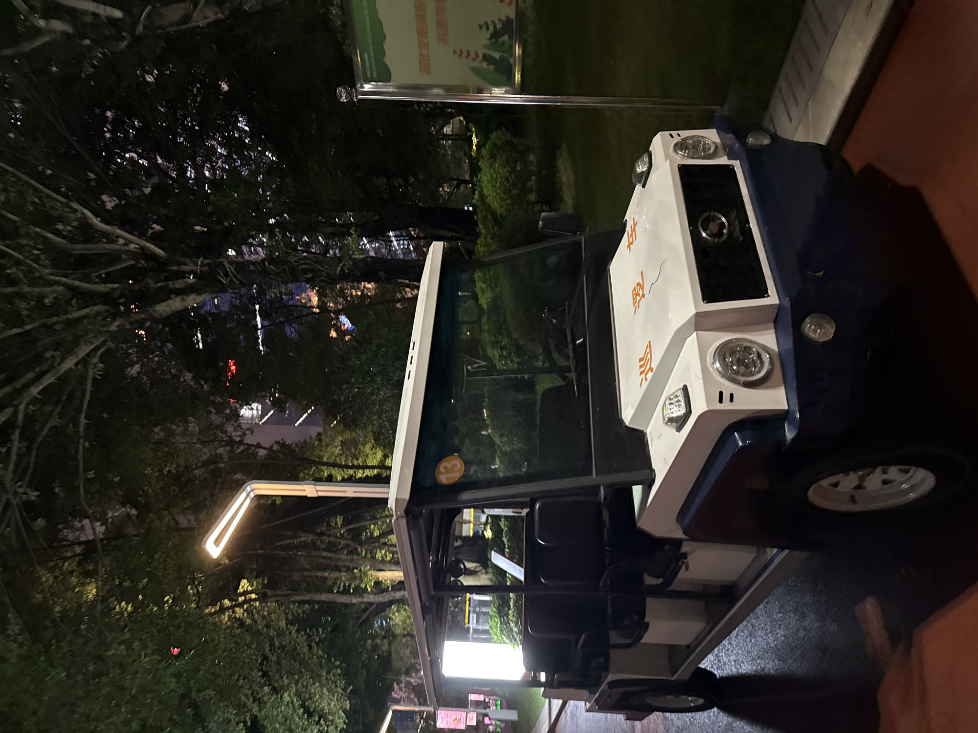 The image shows a white and blue electric vehicle, possibly a golf cart or shuttle, parked on a paved path at night. The vehicle is angled towards the right side of the frame, with its front facing upwards. It has round headlights, turn signals, and a black grille on the front. The side of the vehicle is white with orange Chinese characters printed on it. A blue stripe runs along the lower part of the vehicle's body. The vehicle has black seats visible through its open windows.
Behind the vehicle, a well-lit street lamp with a modern design illuminates the area. The background features dark trees with visible branches and leaves, suggesting an outdoor setting like a park or a campus. In the distance, city lights are visible, indicating an urban environment. The overall lighting suggests it is nighttime. A signpost with some text and graphics is also visible in the background, to the left of the vehicle and behind the street lamp.
The image captures a static scene with no people or obvious activity taking place. The environment appears calm and quiet. The presence of an electric vehicle and the well-maintained path suggest it might be a controlled or managed area. The Chinese characters on the vehicle are the only visible text.