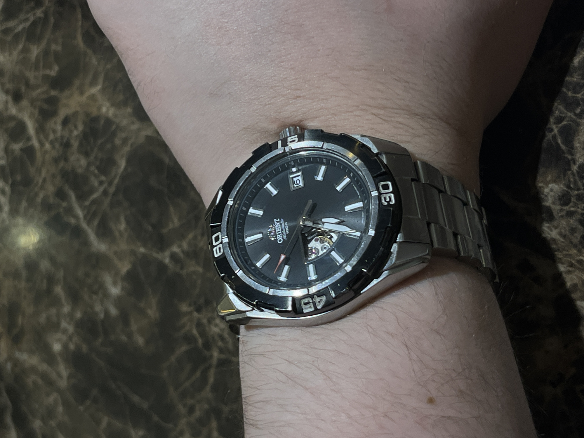 This is a close-up, eye-level photograph of a person's wrist wearing a watch. The watch has a silver metal band and a black face with silver hour markers and hands. The bezel of the watch is also black with silver markings, indicating minutes. A small window on the watch face reveals the internal gears of the automatic movement. The watch is positioned on the person's left wrist, which is resting on a dark, marbled surface. The lighting suggests an indoor setting, possibly with a direct light source creating a soft glow on the watch and skin. The person's skin appears fair and has fine hairs visible. Text on the watch face includes "ORIENT," "Automatic," and "100m." The bezel also features numbers like "60," "30," and "45." The image does not provide clear cues for time of day, weather, or specific emotions, but the focus on the watch suggests an interest in timekeeping or the accessory itself. The marbled surface could be a table or countertop. The location context of Almaty, Kazakhstan, is external information and not directly discernible from the image itself.