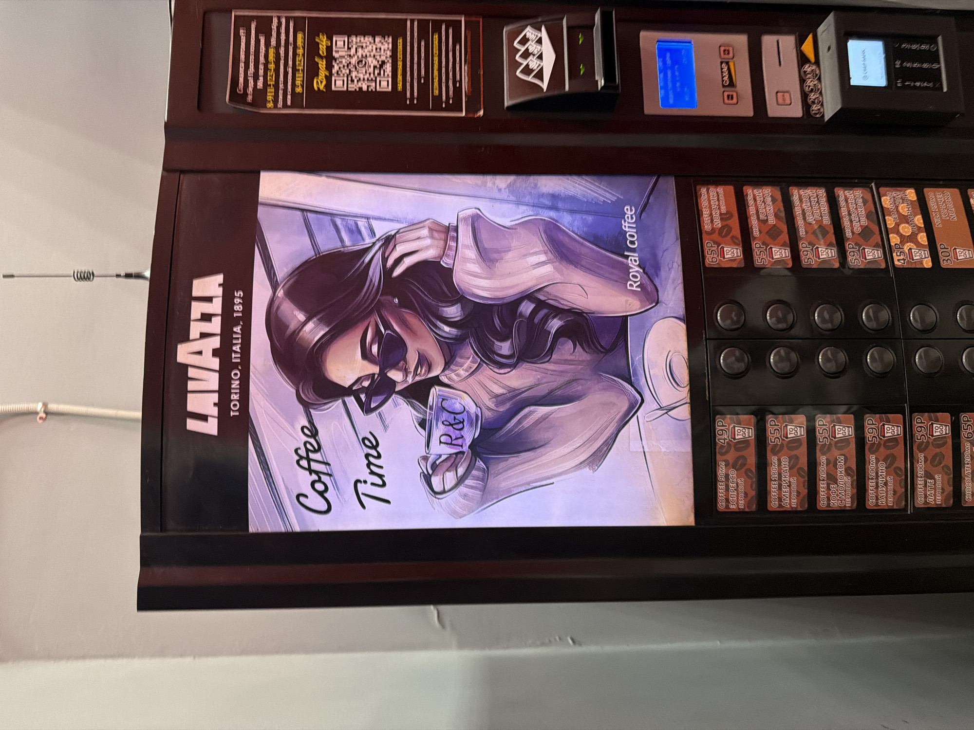 Here is a detailed description of the image:
The image shows a coffee vending machine located in an unknown city in Russia. The machine's front panel features a stylized illustration of a woman wearing sunglasses and holding a coffee cup with the initials "R&C." The machine is branded with "LAVAZZA Torino, Italia, 1895" and promotes "Royal Coffee."
Above the illustration, there's a contact information panel with a QR code and Russian text offering assistance if the machine malfunctions, along with phone numbers "8-911-123-4-999." There are also slots and buttons for payment, along with screens and various selections for different types of coffee.
The coffee selection options include:
* Espresso (49P)
* Moccachino (65P)
* Americano (55P)
* Coffee with milk (55P)
* Cappuccino (59P)
* Latte (59P)
* Chocolate (65P)
* Double hot chocolate (59P)
* Creamy chocolate (59P)
* Lemon tea (45P)
* Milk (30P)
The background is a plain wall, suggesting an indoor location. A white cable is attached to the wall.